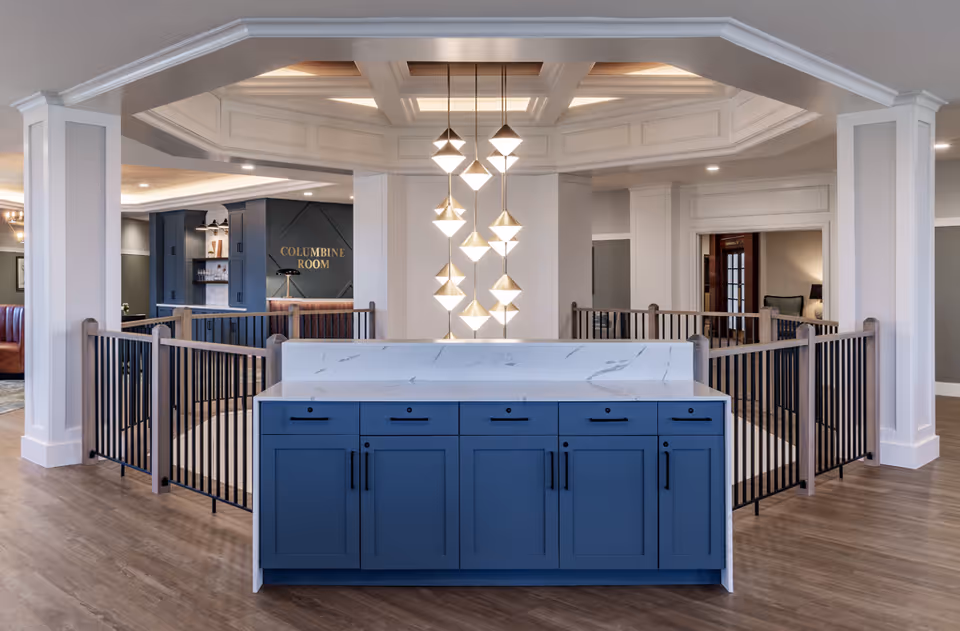 Interior view of a senior living facility featuring a modern blue cabinet with a white marble countertop in the foreground. Above the cabinet hangs a contemporary chandelier with geometric light fixtures. The space is open with white columns and railings, and in the background, there is a sign for the Columbine Room and seating areas with leather chairs.