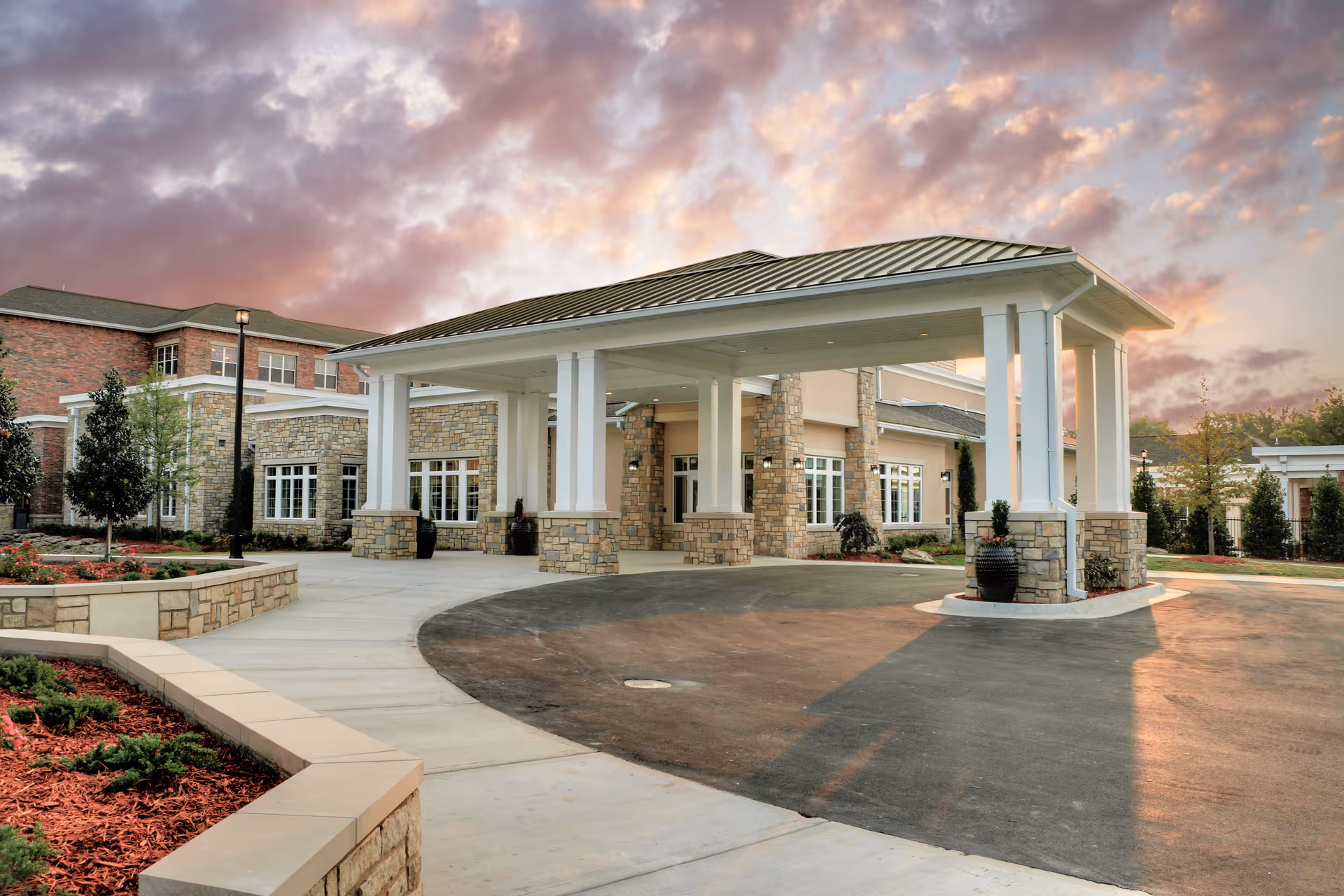 Entrance of a senior living facility with a covered drop-off area supported by white columns and stone bases, surrounded by landscaped gardens and a paved driveway under a colorful sky at sunset.