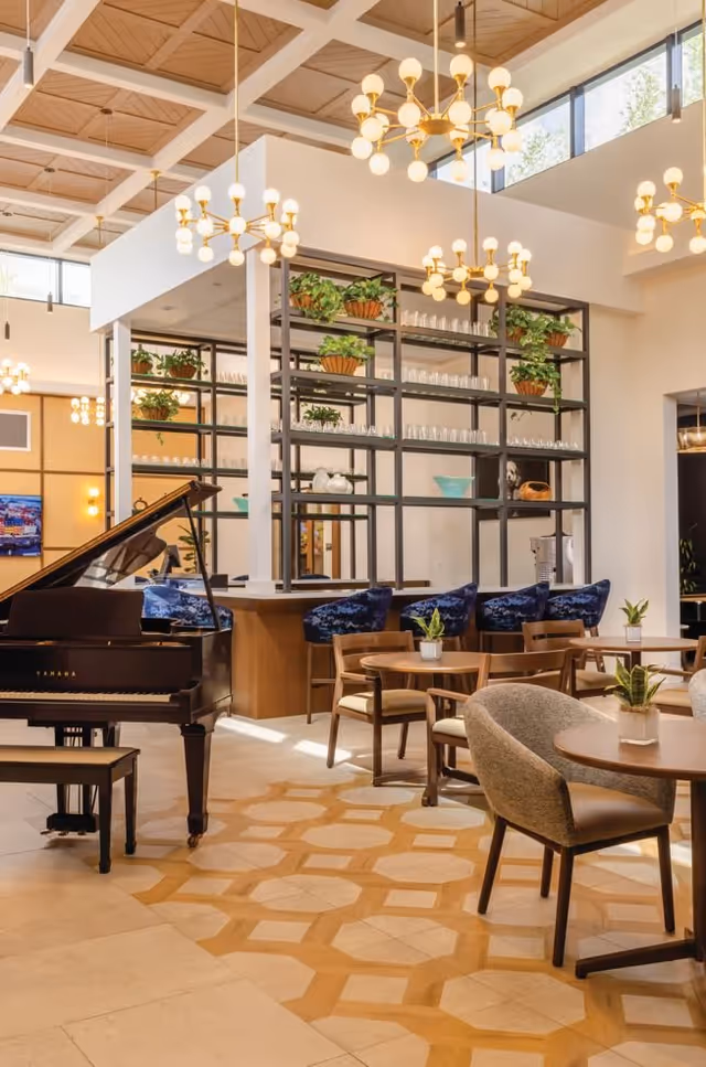 A bright and spacious lounge area with a grand piano, round wooden tables with chairs, and a bar counter with blue cushioned stools. The room features modern chandeliers, large windows, and decorative plants on shelves behind the bar.