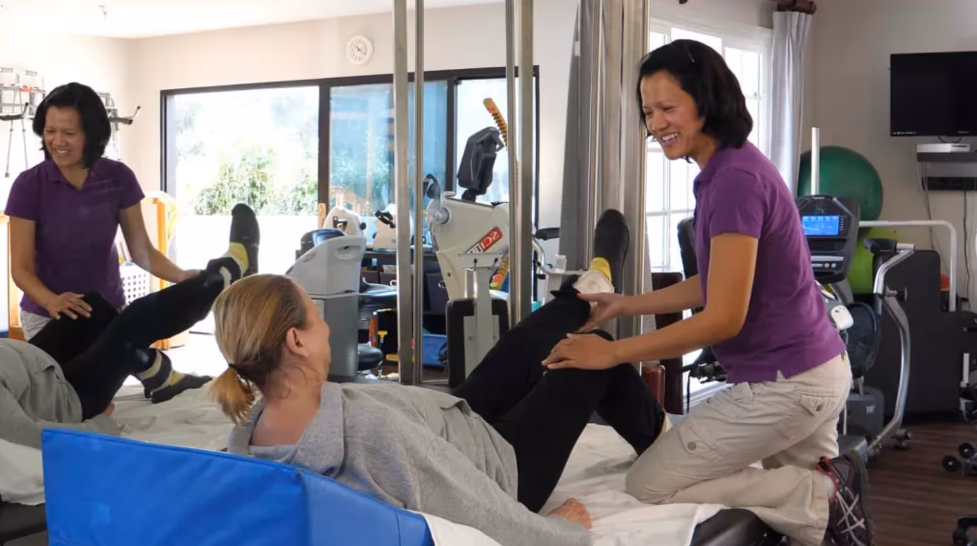 A physical therapist in a purple shirt assists a woman lying on a treatment table with leg exercises in a bright therapy room equipped with exercise machines and mirrors.