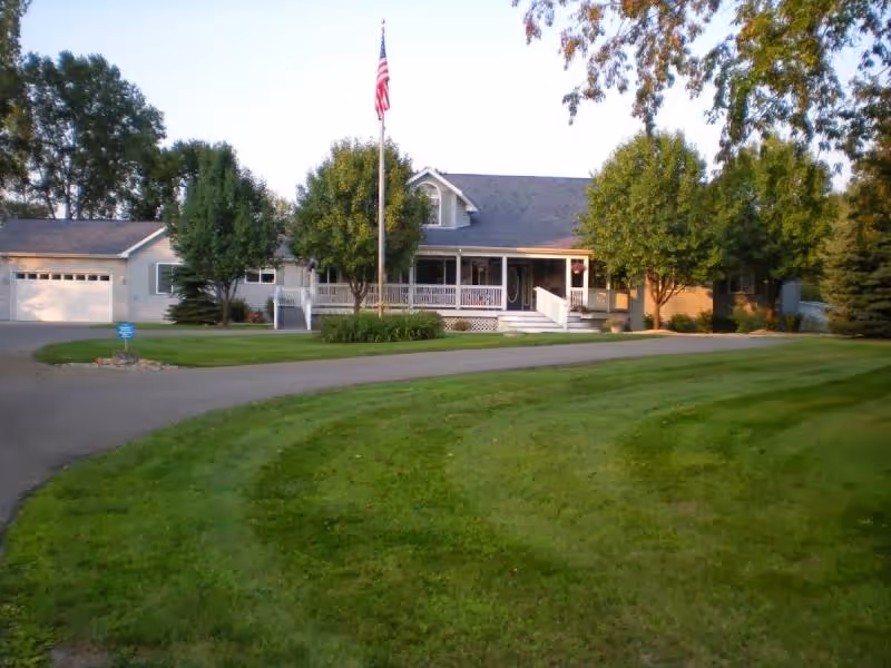 A single-story residential building with a large front porch, surrounded by green trees and a well-maintained lawn. An American flag is displayed on a flagpole in front of the house. The driveway curves around the lawn leading to a garage attached to the house.