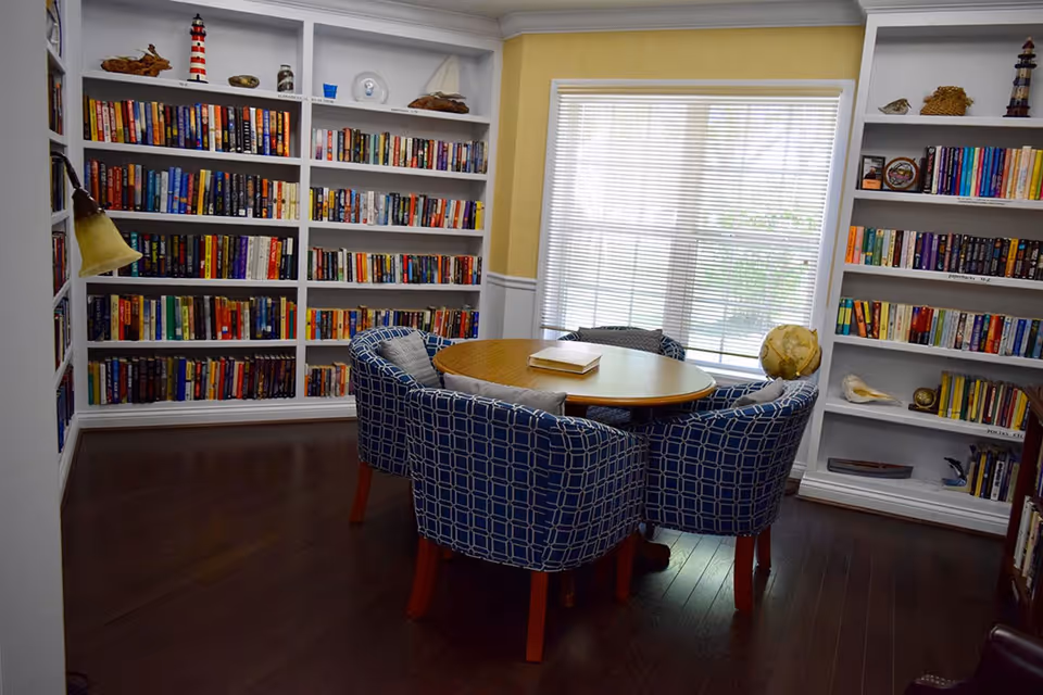 A cozy reading room with built-in white bookshelves filled with books and decorative items. In the center, there is a round wooden table surrounded by four blue patterned armchairs. A large window with blinds allows natural light to brighten the room.