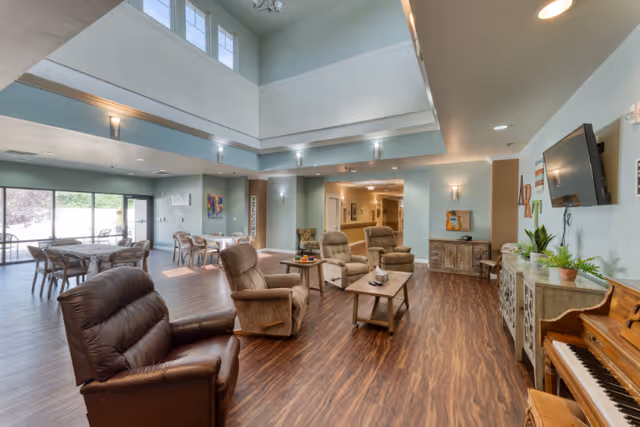 A spacious and well-lit common area in a senior living facility featuring comfortable recliner chairs arranged around a coffee table, a piano on the right side, a wall-mounted TV, and several tables and chairs near large windows letting in natural light. The room has high ceilings with upper windows and soft wall lighting.