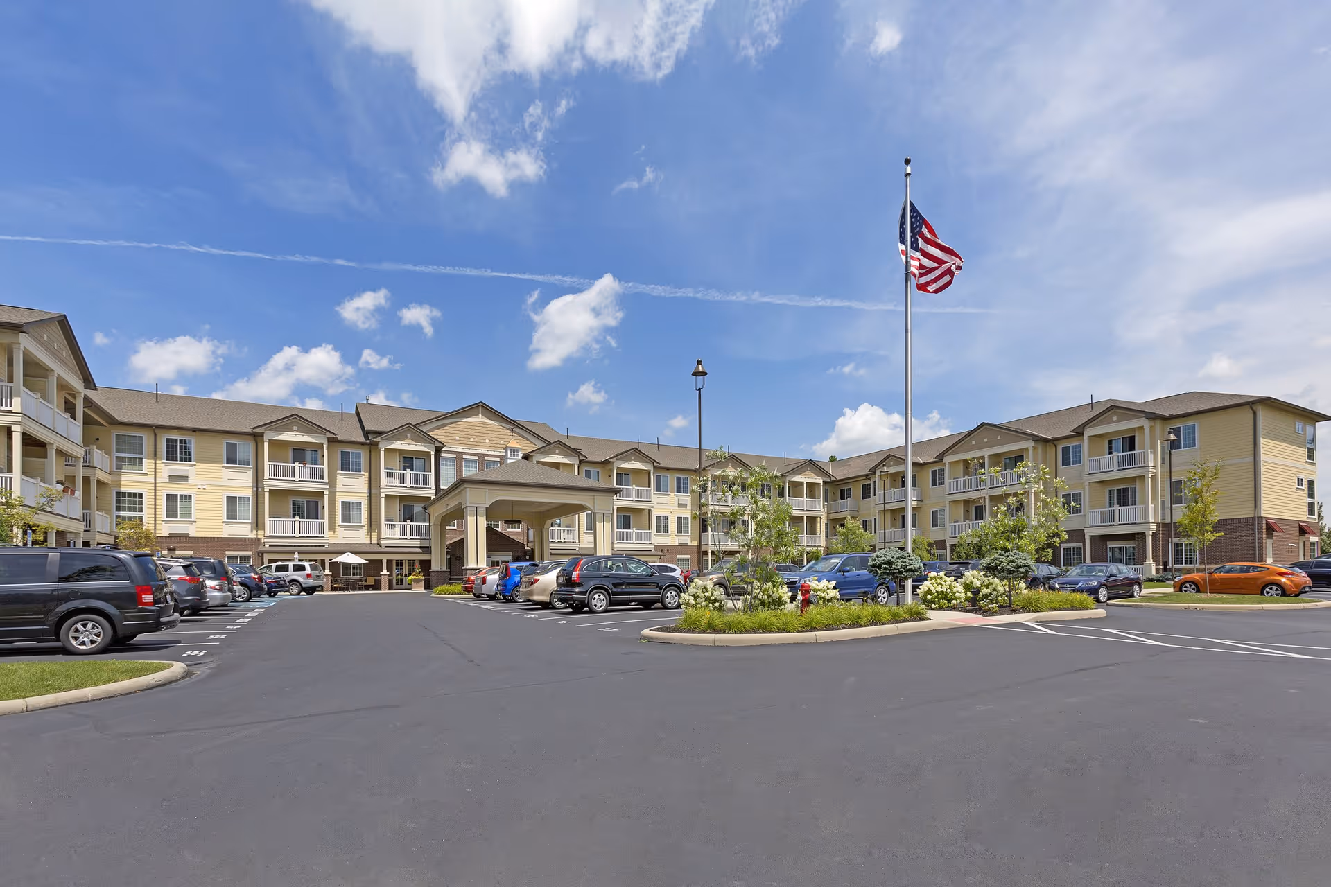 Exterior view of a three-story senior living facility building with beige siding and balconies. There is a large parking lot in front with several parked cars and a tall flagpole displaying the American flag. The sky is partly cloudy with blue patches.