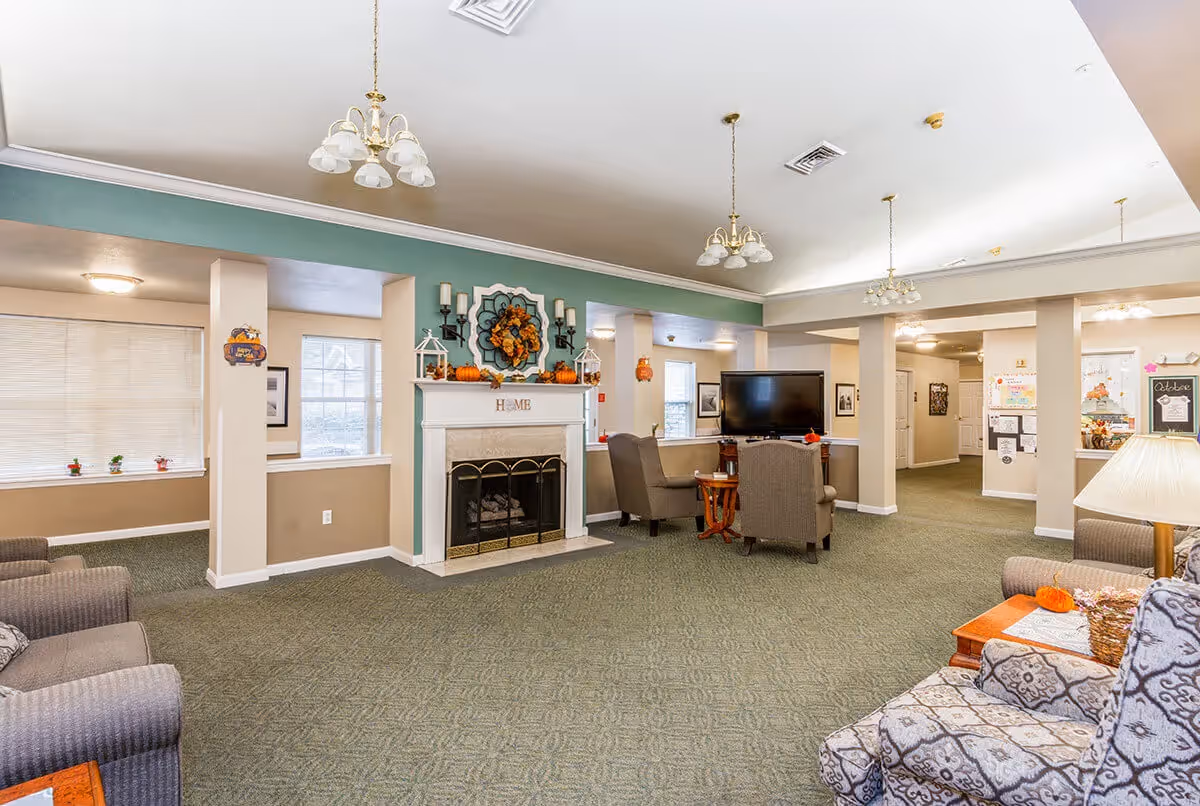A spacious living room area in a senior living facility with a green carpet and beige walls. The room features a white fireplace decorated with autumn-themed ornaments and the word 'HOME' on the mantel. There are several comfortable armchairs arranged around a flat-screen TV. The ceiling has multiple hanging light fixtures, and there are windows letting in natural light. The room has a cozy and welcoming atmosphere with seasonal decorations.
