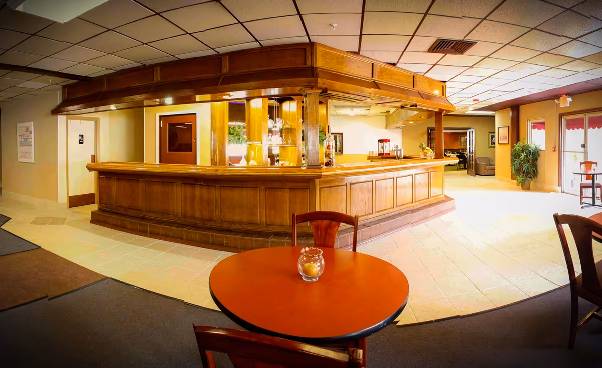 Interior view of a senior living community common area featuring a large wooden reception or bar counter with glass shelves and decorative items. In the foreground, there is a round wooden table with two chairs and a small glass candle holder. The room has tiled flooring, a drop ceiling with fluorescent lights, and a doorway leading to other rooms. There are plants and framed pictures on the walls, and large windows with red awnings letting in natural light.