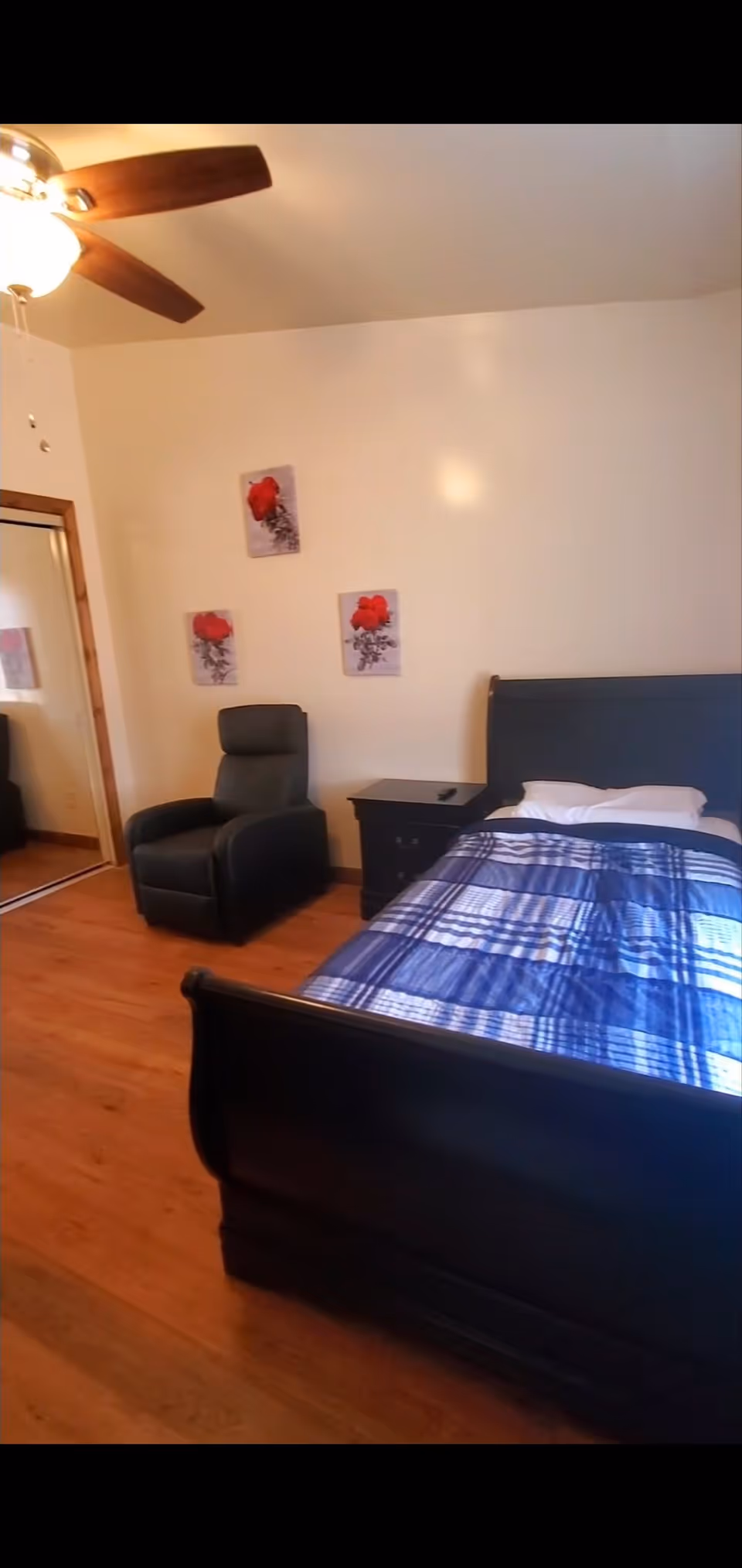 A bedroom with a dark wooden bed frame and a blue plaid bedspread. There is a black armchair next to a black nightstand with a remote control on it. Three wall art pieces featuring red flowers are hung on the beige wall above the armchair. A ceiling fan with a light is visible on the ceiling, and a mirrored closet door is on the left side of the room. The floor is wooden.
