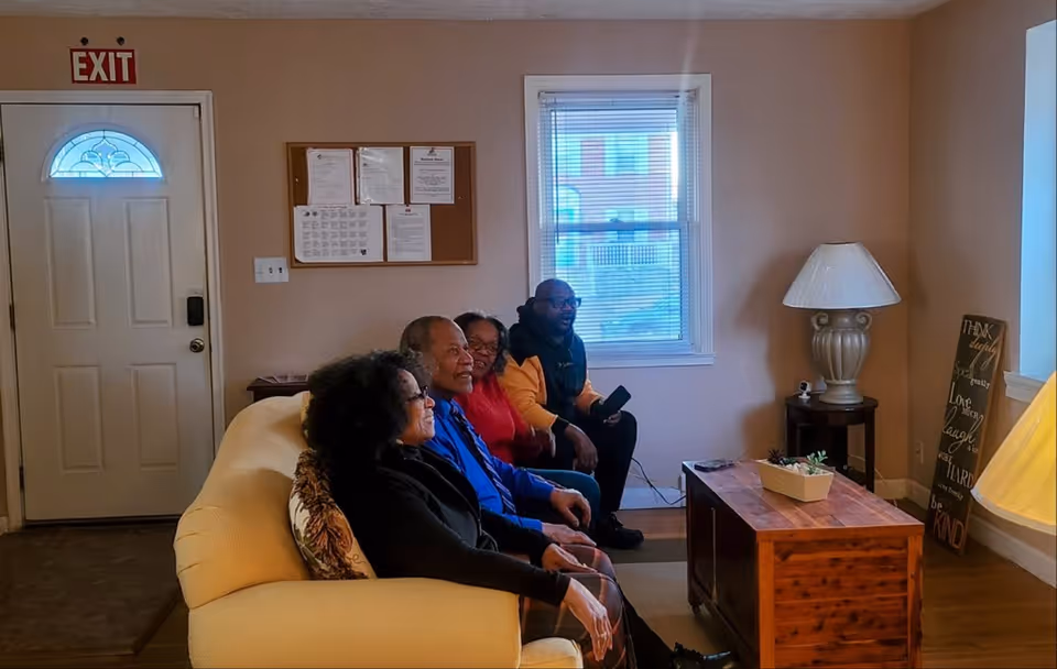 Four people sitting on a beige couch in a living room with a wooden coffee table, a lamp on a side table, a window with blinds, and a door with an exit sign above it.
