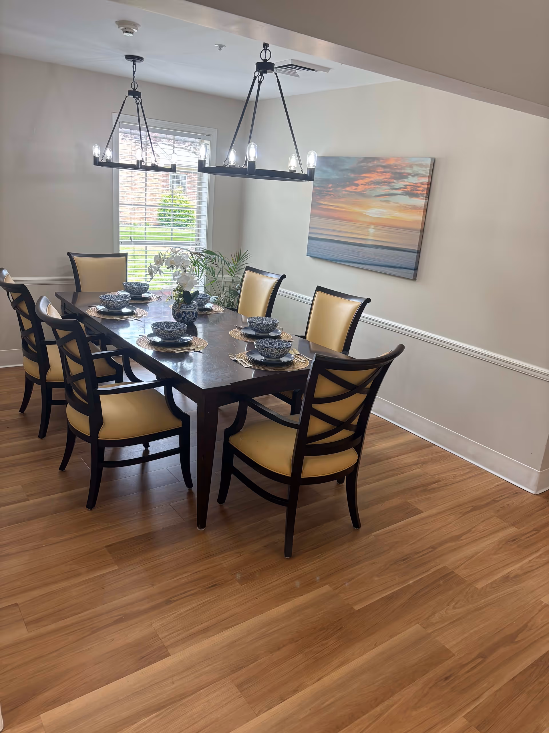 A dining room with a dark wooden rectangular table set for six people with matching chairs that have yellow cushions. The table is decorated with blue and white bowls and plates, and a small floral centerpiece. There is a window with white blinds letting in natural light, a painting of a sunset over the ocean on the wall, and two modern black chandeliers hanging from the ceiling. The floor is wooden.