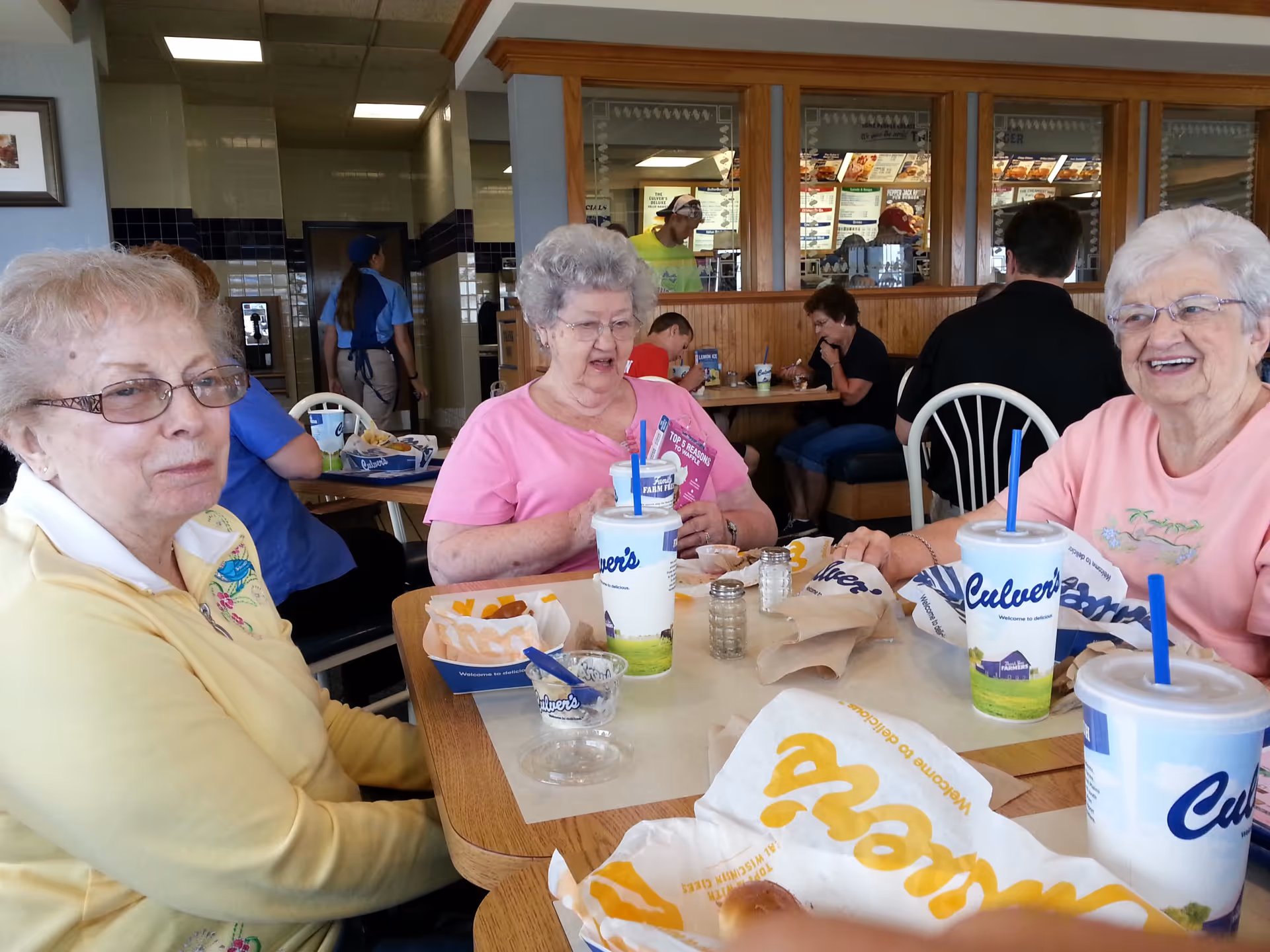 Three elderly women sitting around a table at a Culver's restaurant, enjoying food and drinks. The table has wrappers, cups, and food items. Other diners and restaurant staff are visible in the background.