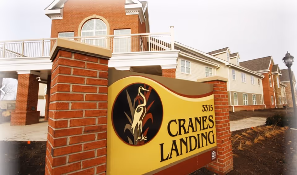 Exterior view of Cranes Landing facility showing a large yellow and brown sign with a crane bird logo and the address number 3315, set between two brick pillars. The building behind the sign is a multi-story structure with brick and beige siding, multiple windows, and a balcony with white railings.