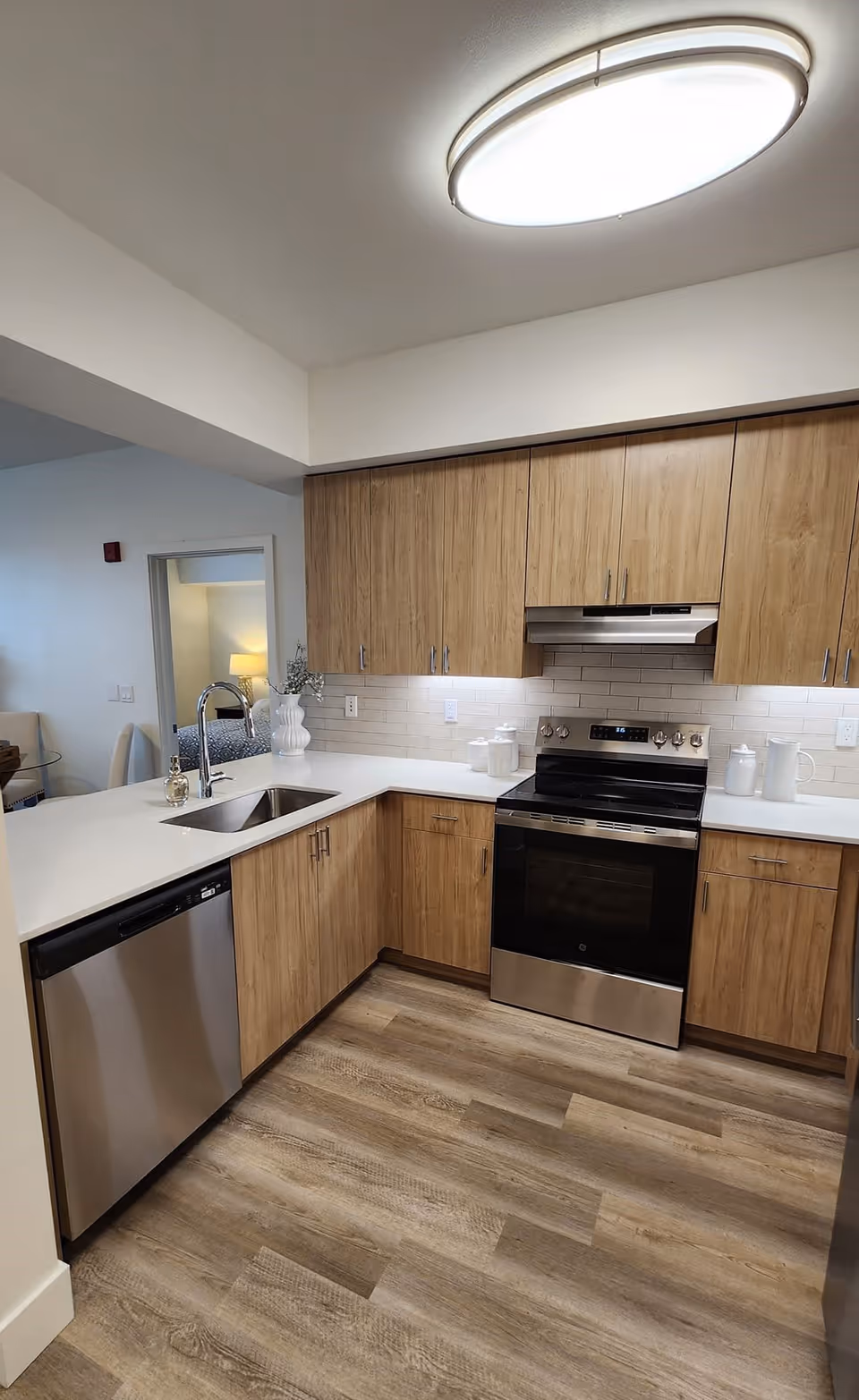 Modern kitchen with light wood cabinets, white countertops, stainless steel dishwasher and oven, and a white subway tile backsplash. The kitchen has a sink with a curved faucet and under-cabinet lighting. The floor is wood-patterned, and there is a circular ceiling light fixture.