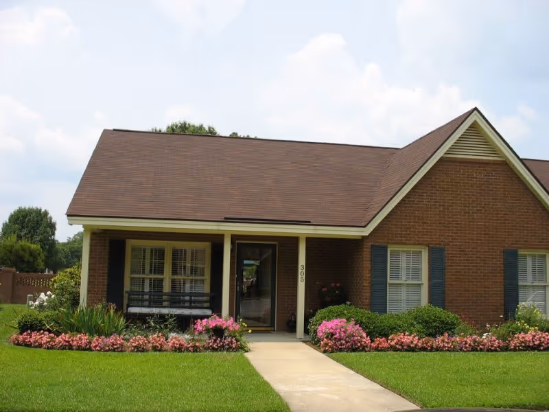 Single-story brick building with a brown shingled roof, white trim, and blue shutters on the windows. There is a concrete walkway leading to a glass front door with the number 305 beside it. The front yard is well-maintained with green grass, bushes, and pink flowers lining the walkway and the front of the building. The sky is partly cloudy.