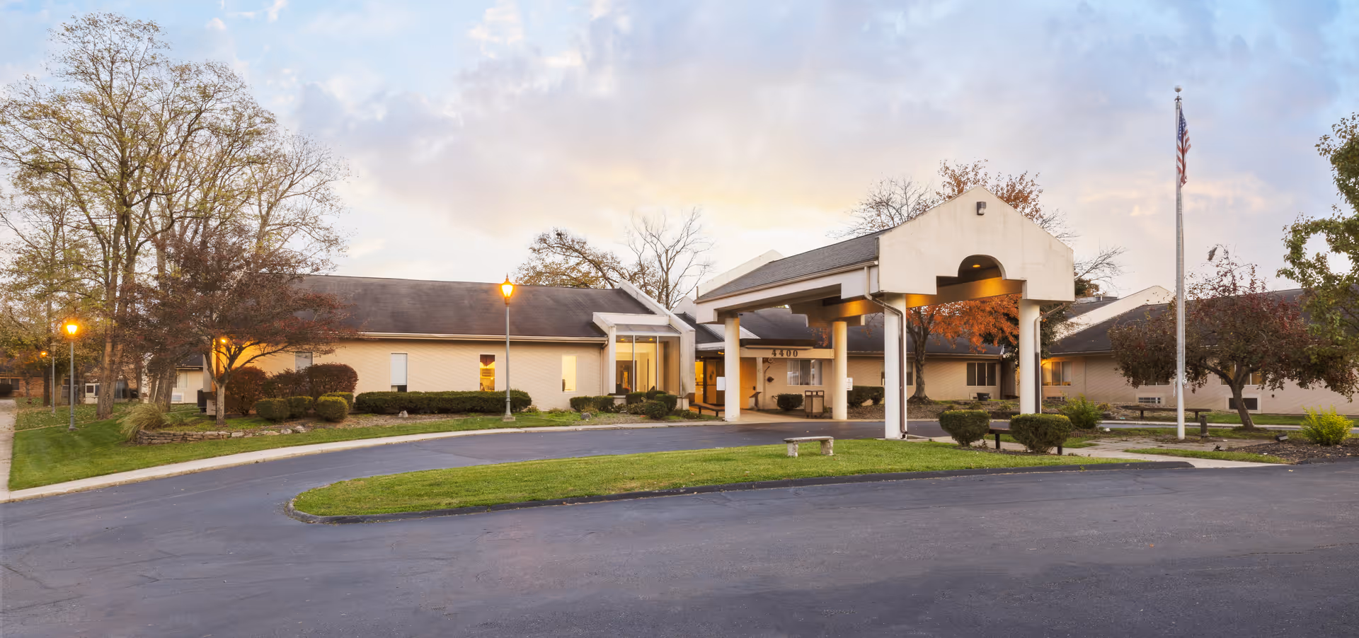 Front exterior of a senior living facility showing a covered entrance/porte-cochère, circular driveway, landscaped lawn, and flagpole at dusk.