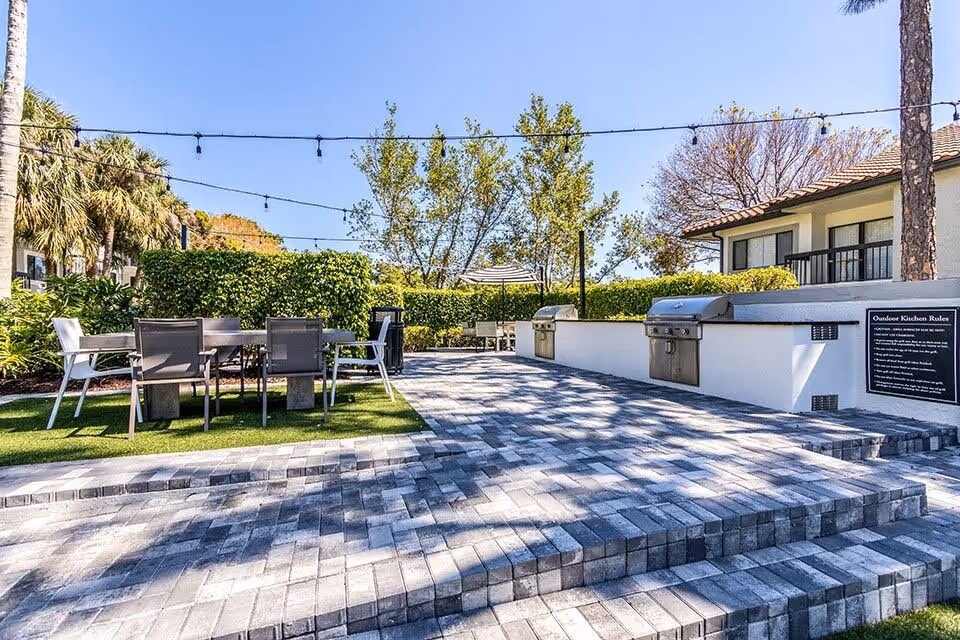 Outdoor communal patio with dining tables and chairs, built-in stainless steel grills, string lights, and paver steps.