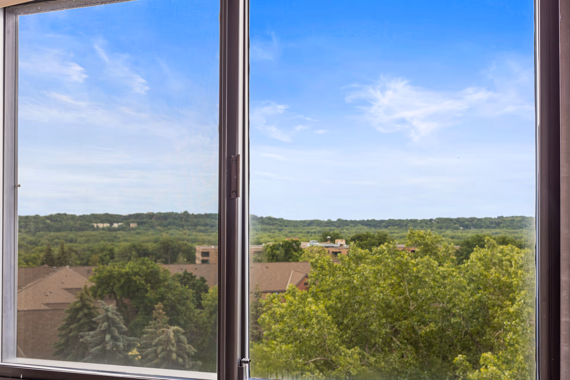 View through a large window showing a landscape with green trees, rooftops of buildings, and a blue sky with some clouds.