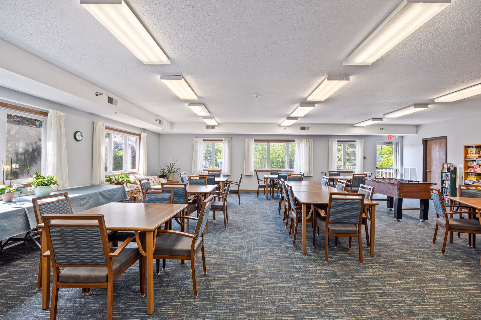 A spacious, well-lit common room with multiple wooden tables and chairs arranged neatly. Large windows with white curtains allow natural light to fill the room. There are plants on a side table covered with a blue tablecloth, a foosball table, and shelves with board games or books. The ceiling has multiple rectangular fluorescent lights.