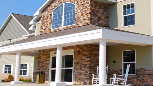 Exterior view of a senior living facility with a stone and stucco facade, large windows, a covered entrance supported by white columns, and two white rocking chairs on the porch.