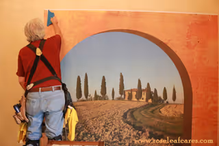 A person wearing a red shirt and blue jeans is cleaning or installing a large wall mural depicting a scenic countryside view with a house, trees, and a curved path under a blue sky. The mural is framed by an arch painted on the wall.