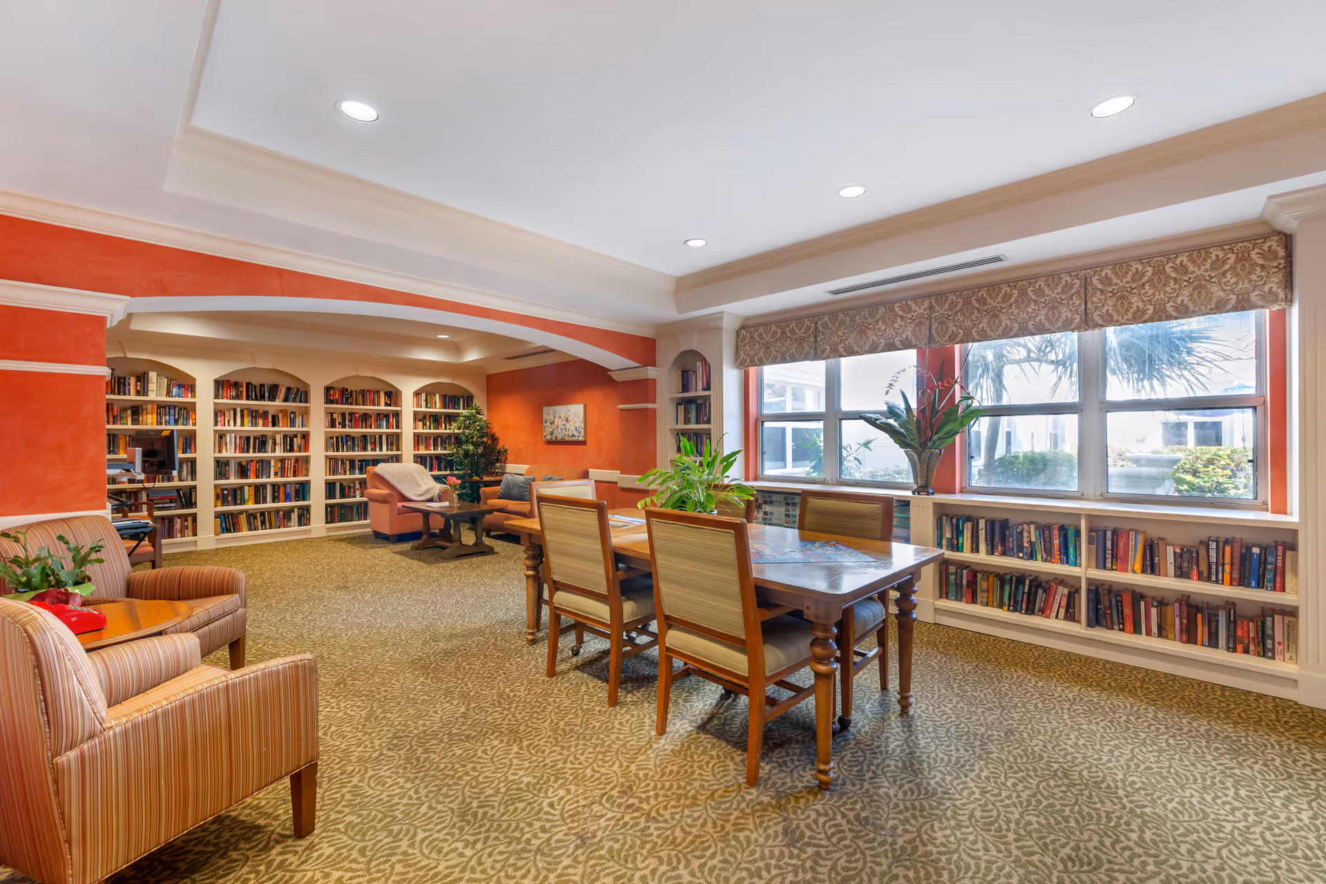 Bright communal lounge and library area with bookshelves, seating, and a table by large windows.