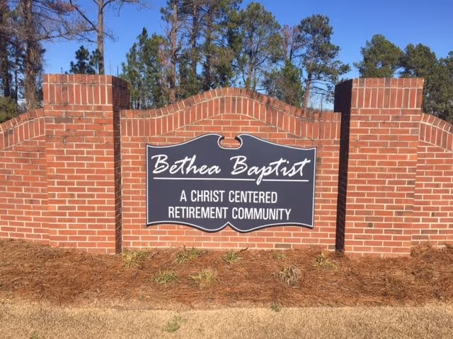 A brick sign at the entrance of Bethea Baptist, a Christ centered retirement community, with trees and a clear blue sky in the background.