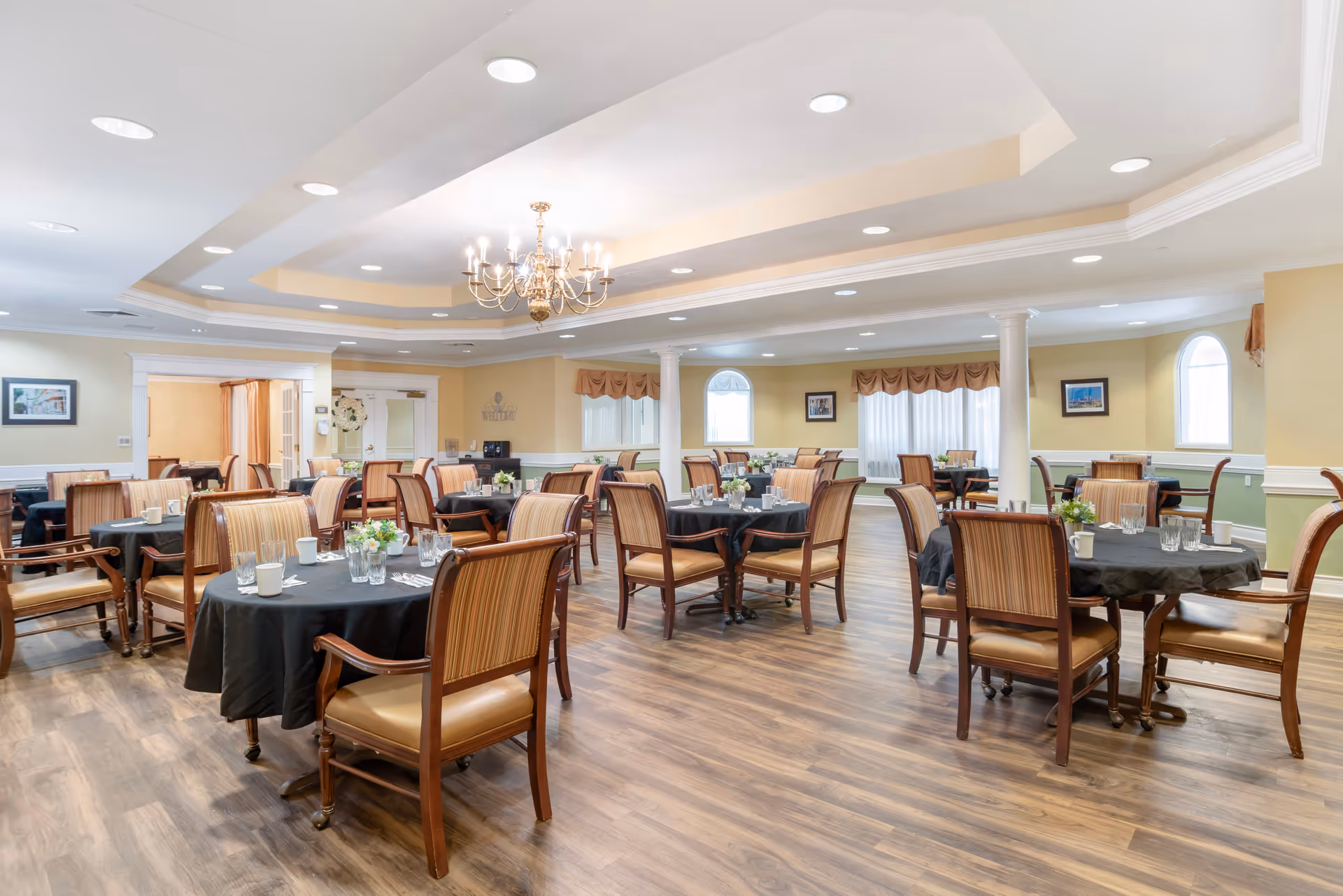A spacious dining room with multiple round tables covered with black tablecloths, each surrounded by wooden chairs with striped upholstery. The room features wood flooring, soft yellow walls with white trim, decorative columns, and a chandelier hanging from a recessed ceiling. Windows with curtains and framed pictures adorn the walls.