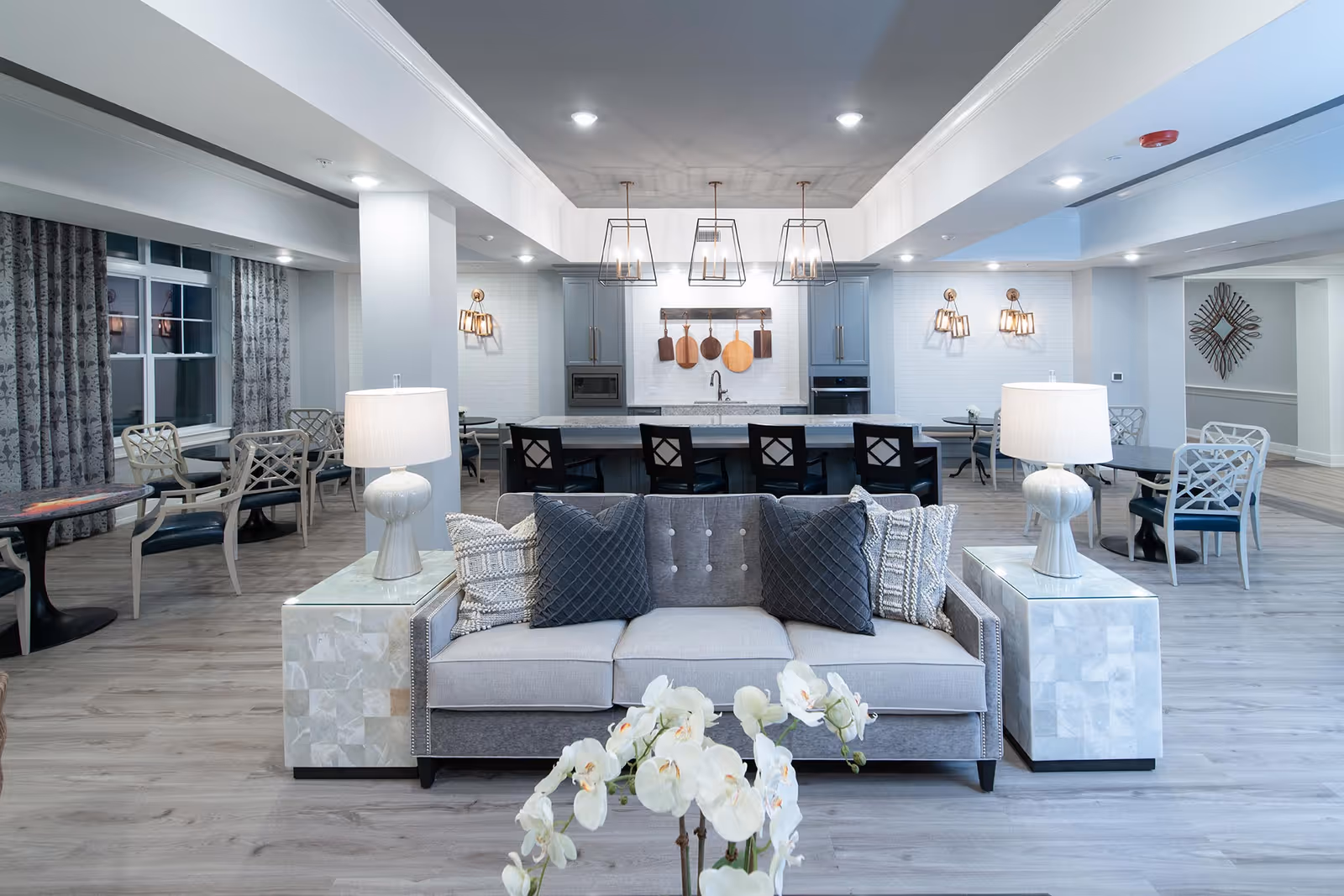 A modern senior living common area featuring a gray sofa with decorative pillows in the foreground, two side tables with white lamps, and a kitchen area in the background with hanging pendant lights and wooden cooking utensils on the wall. There are multiple seating areas with tables and chairs along the sides, light wood flooring, and large windows with patterned curtains.