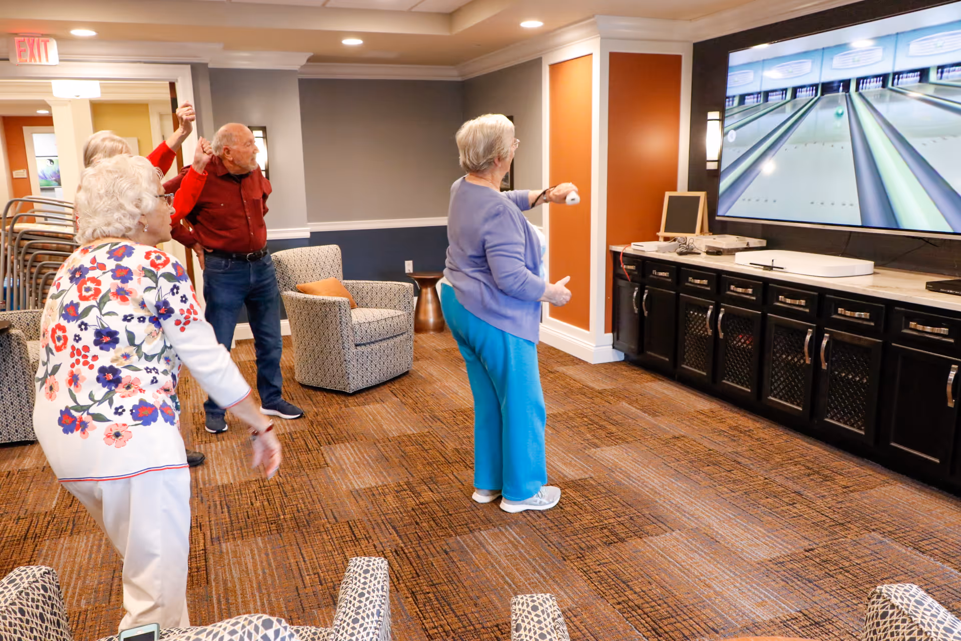 A group of elderly people playing a virtual bowling game on a large screen in a comfortable common area with patterned carpet, armchairs, and warm lighting.