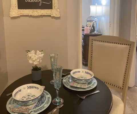 A small round dining table set for two with floral patterned plates, bowls, and cloth napkins. Two tall greenish drinking glasses are placed on the table along with silverware. A small potted white orchid is also on the table. Behind the table is a beige upholstered chair with nailhead trim. In the background, a lamp on a wooden side table and a framed chalkboard with the word 'blessing' written on it are visible.