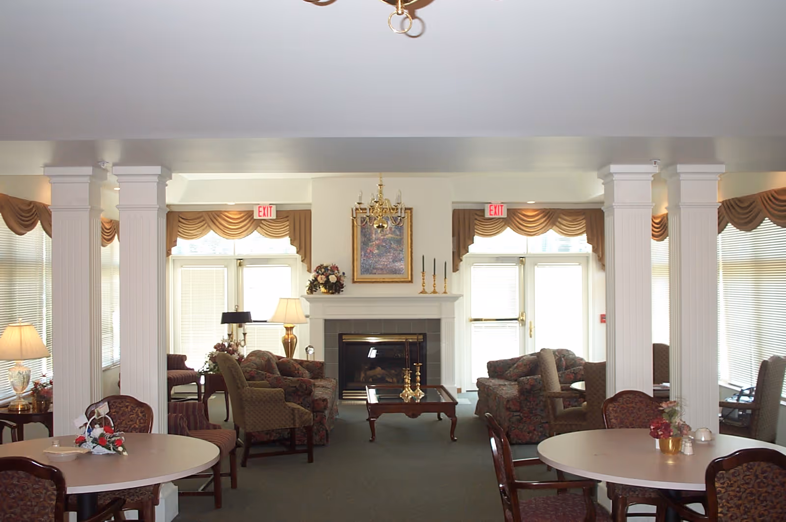 A spacious and well-lit common area with round tables and chairs in the foreground, and several upholstered armchairs and sofas arranged around a central fireplace. The room features large windows with blinds and valances, white columns, table lamps, floral arrangements, and a chandelier hanging from the ceiling.