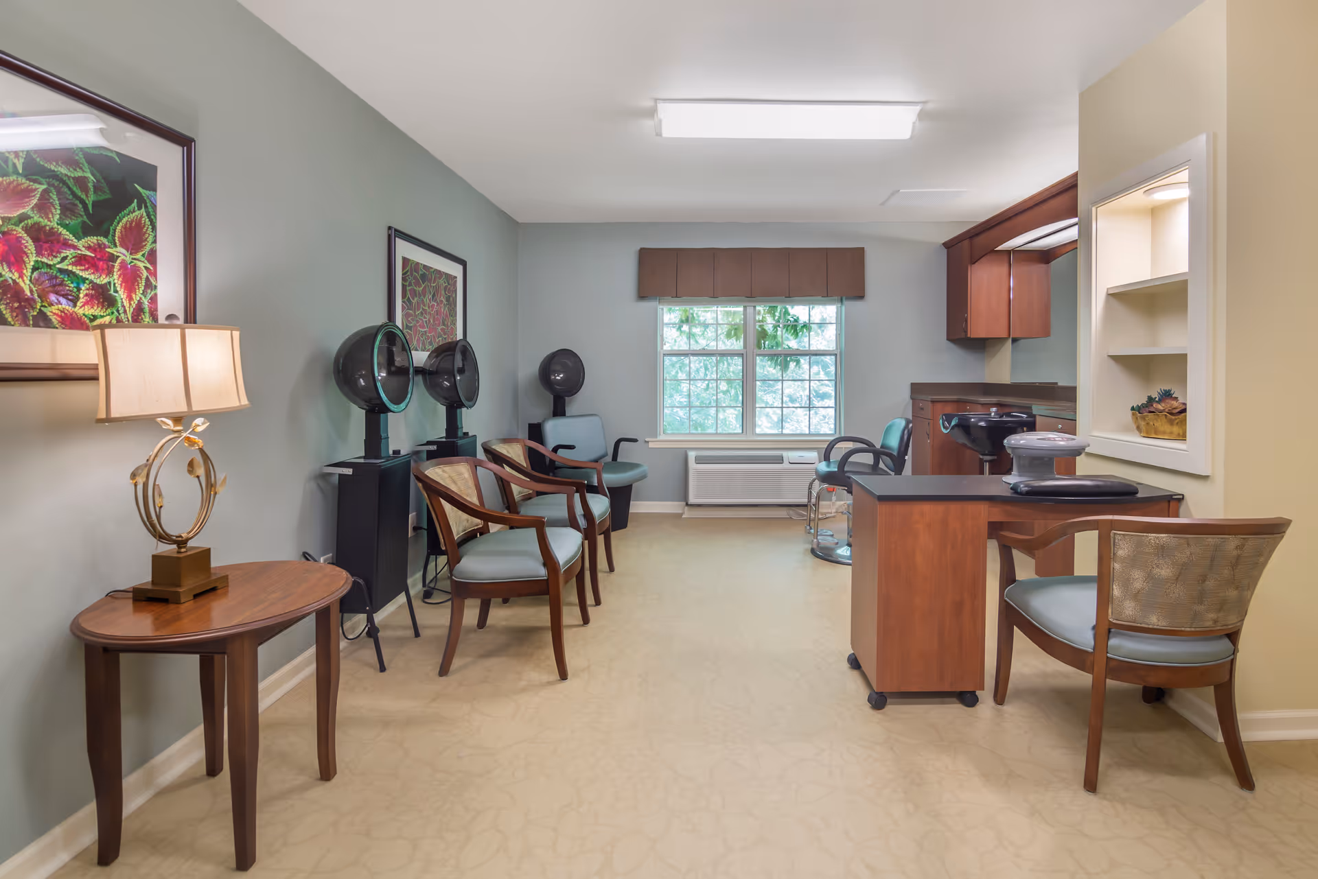 Interior view of a hair salon area in a senior living facility with multiple hair drying stations, chairs, a small table with a lamp, framed artwork on the walls, and a window with a brown valance letting in natural light.
