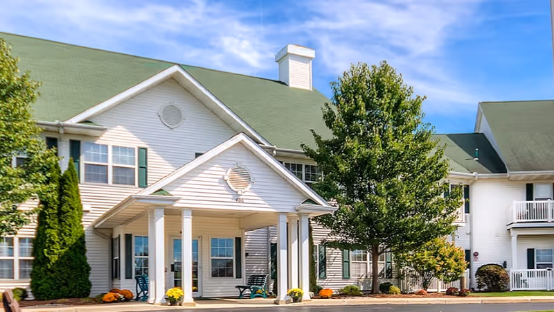 Exterior view of a two-story senior living facility with white siding and a green roof. The entrance features a covered porch with white columns, and there are trees and shrubs around the building under a partly cloudy blue sky.