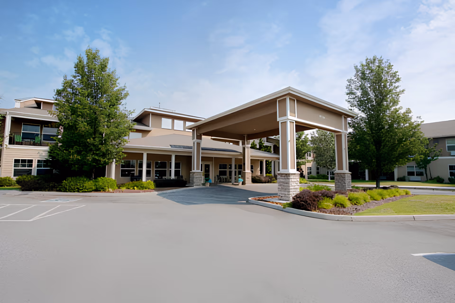 Exterior view of Wenatchee Senior Living by Cogir showing the main entrance with a large covered driveway, surrounded by trees, shrubs, and a well-maintained lawn under a partly cloudy sky.