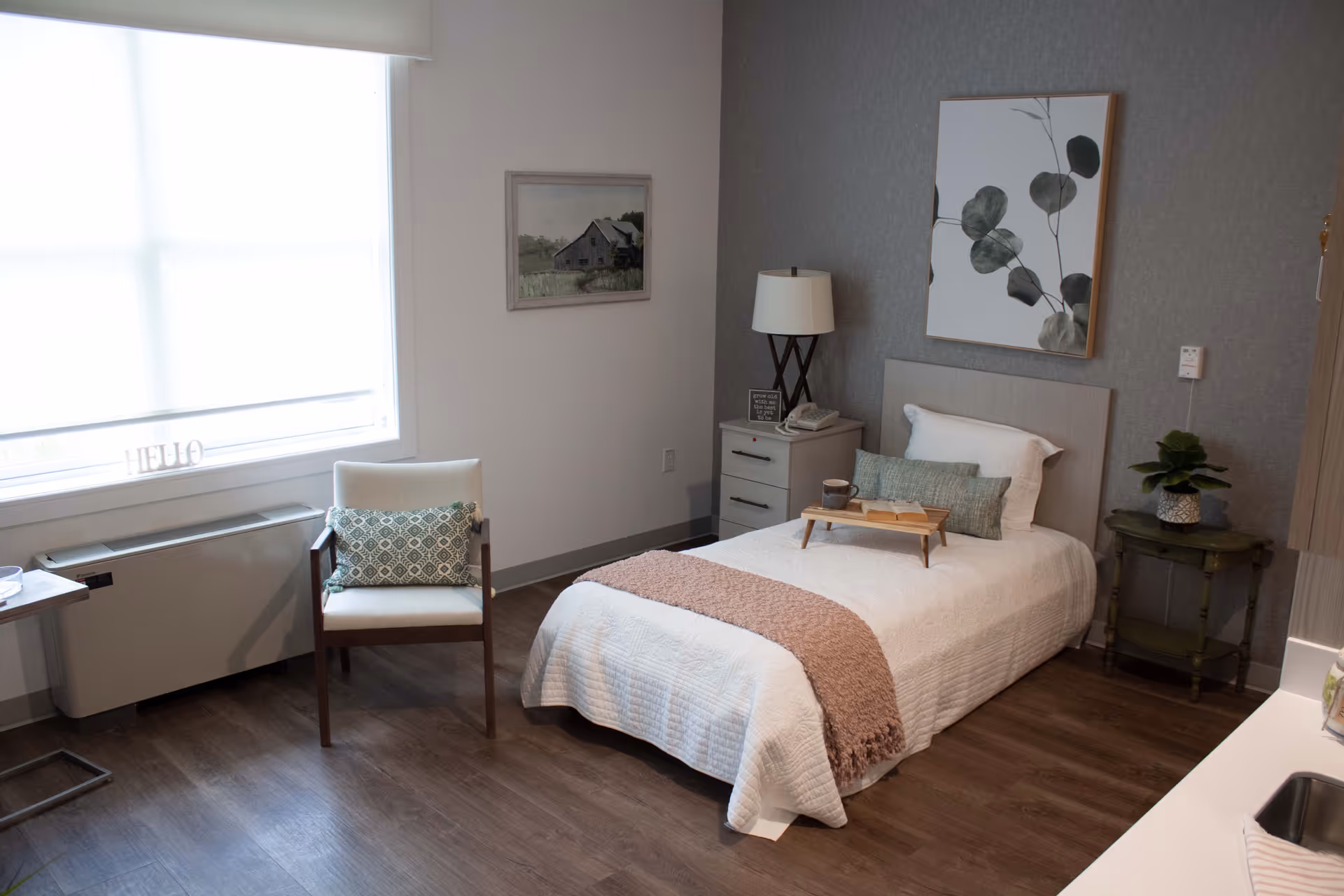 A neatly arranged bedroom in an assisted living facility featuring a single bed with white bedding and a pink throw blanket. There is a small wooden tray on the bed holding a cup and an open book. Next to the bed is a nightstand with a lamp, a phone, and a small decorative sign. A chair with a patterned cushion is placed near a large window with a white blind. The room has wooden flooring, a framed picture on the wall, and a small green side table with a potted plant.