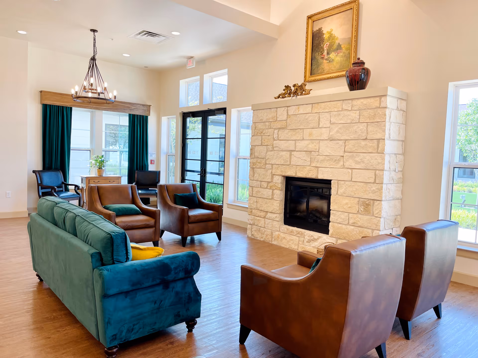 A bright and spacious living room area with a stone fireplace, a green velvet sofa, two brown leather armchairs, and two additional brown chairs near a window with green curtains. A chandelier hangs from the ceiling, and a framed painting and decorative items are placed on the fireplace mantel. Large windows allow natural light to fill the room.