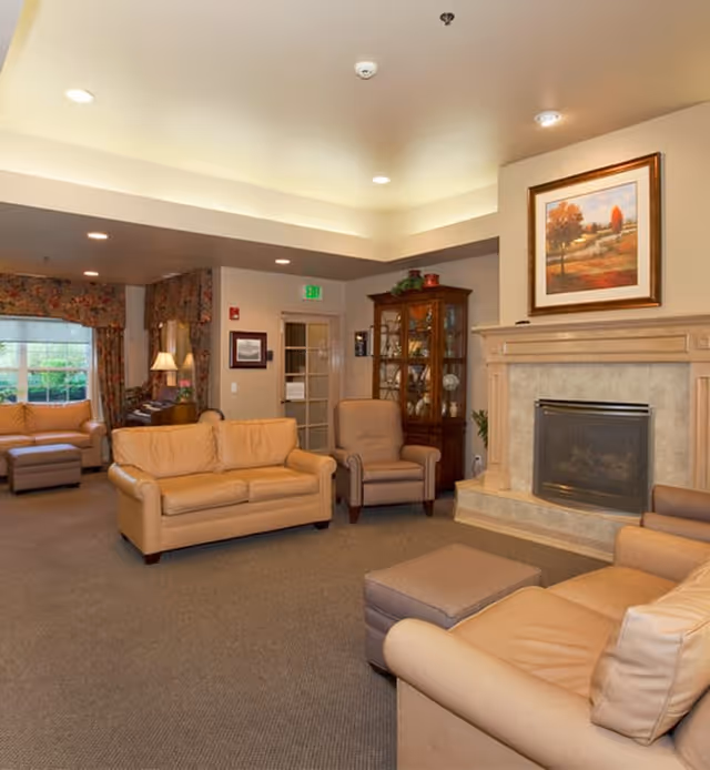 A cozy living room area in a senior living facility featuring beige leather sofas and armchairs arranged around a stone fireplace. A wooden cabinet with glass doors displays decorative items, and a framed painting of an autumn landscape hangs above the fireplace. The room has carpeted flooring, recessed lighting, and floral curtains on the windows.