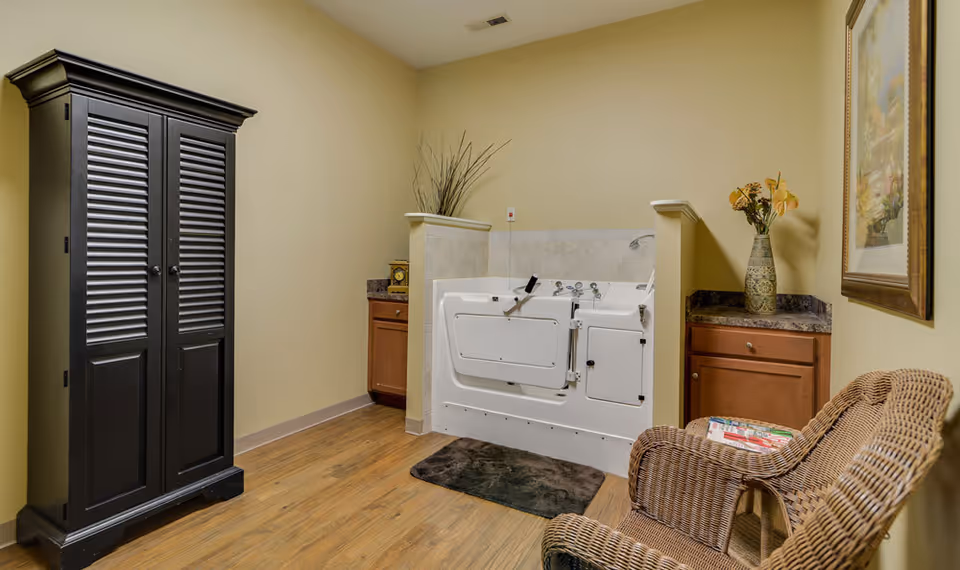 A room with a walk-in bathtub centered against the back wall, flanked by two wooden cabinets with dark countertops. To the left is a tall black cabinet, and to the right is a wicker chair with magazines on it. The walls are painted beige and the floor is wood. A framed picture and a vase with flowers are on the right side.