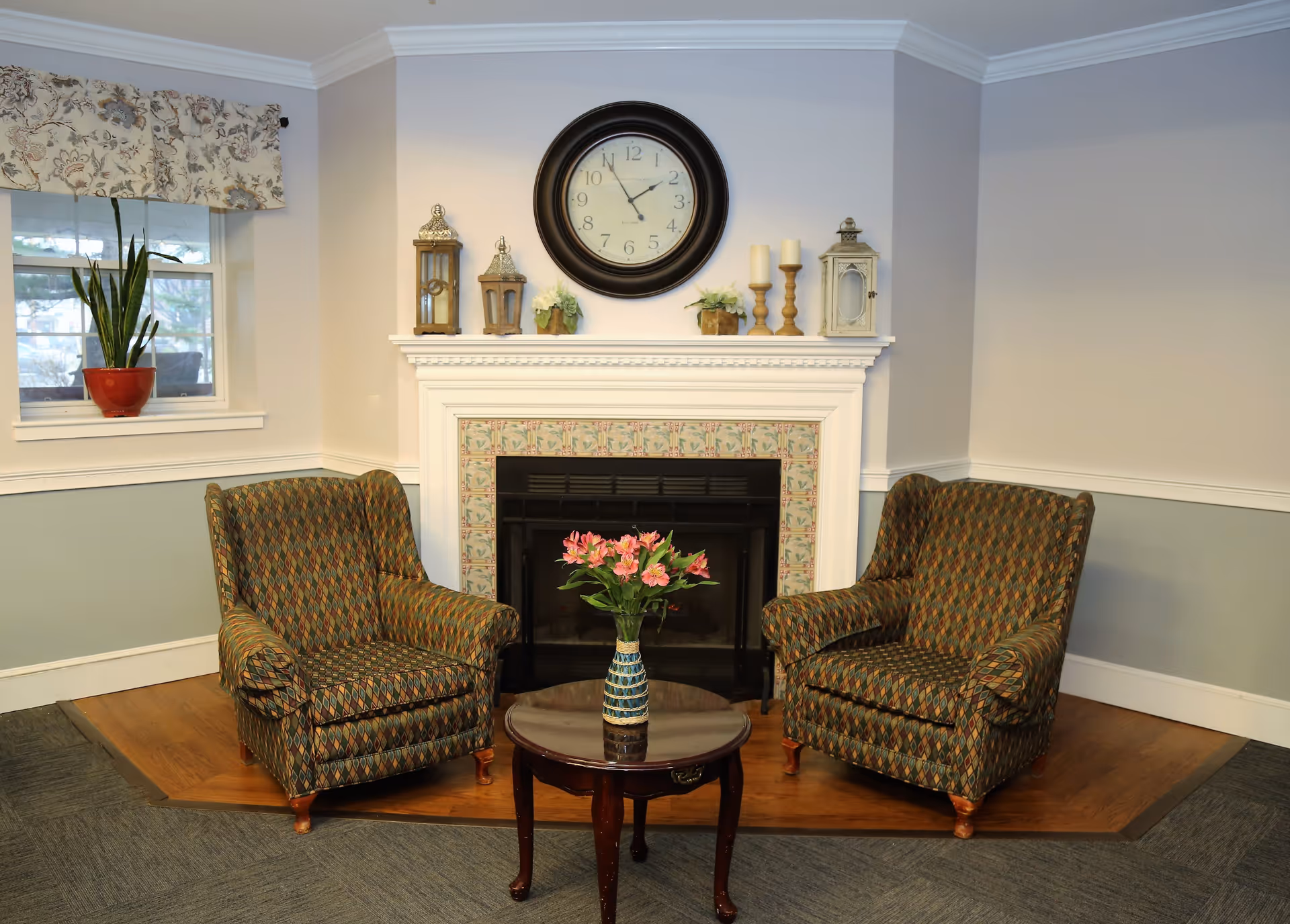 A cozy sitting area with two patterned armchairs facing each other, a small round wooden table with a vase of pink flowers between them, and a white fireplace with decorative tiles and various candle holders and plants on the mantel. A large round clock hangs above the fireplace, and a window with a floral valance and a potted plant is visible on the left side.