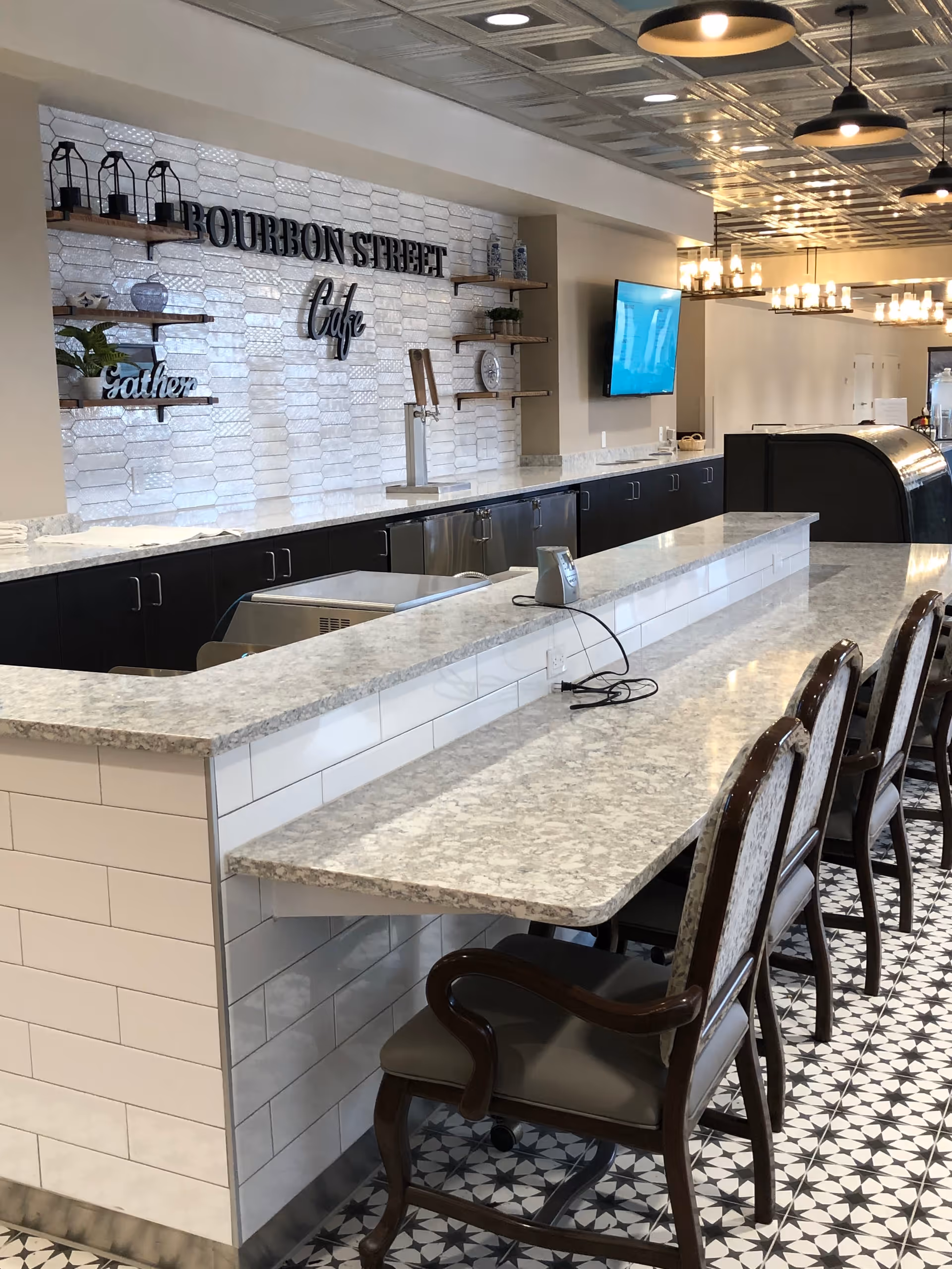 Interior view of a cafe area named Bourbon Street Cafe with a long marble countertop and several wooden chairs with armrests. The wall behind the counter features white textured tiles, wooden shelves with decorative items, and a mounted flat-screen TV. The floor has a patterned tile design and there are modern pendant lights hanging from the ceiling.
