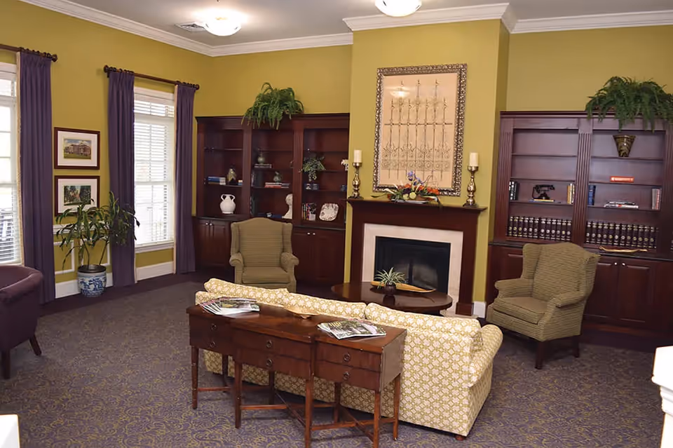 A cozy living room with a patterned yellow sofa facing a fireplace. Two green armchairs flank the fireplace, which has a decorative framed artwork above it and two candle holders on the mantel. Behind the sofa is a wooden console table with magazines on it. The room features built-in dark wood bookshelves with various decorative items and plants. Large windows with purple curtains allow natural light into the room, and there are additional plants placed around the space.