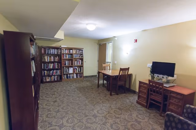 A quiet room with carpeted floor and yellow walls featuring a wooden bookshelf filled with books, a wooden table with two chairs, and a wooden desk with a computer and chair. The room is softly lit with ceiling and wall lights.