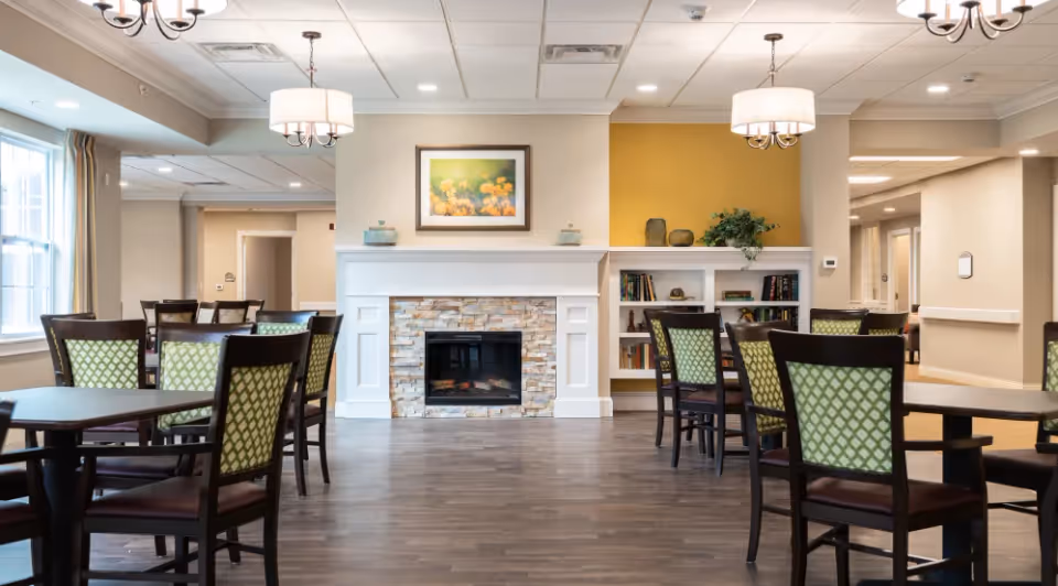 Bright common dining room with multiple tables and green-patterned chairs facing a decorative fireplace and built-in bookshelves.