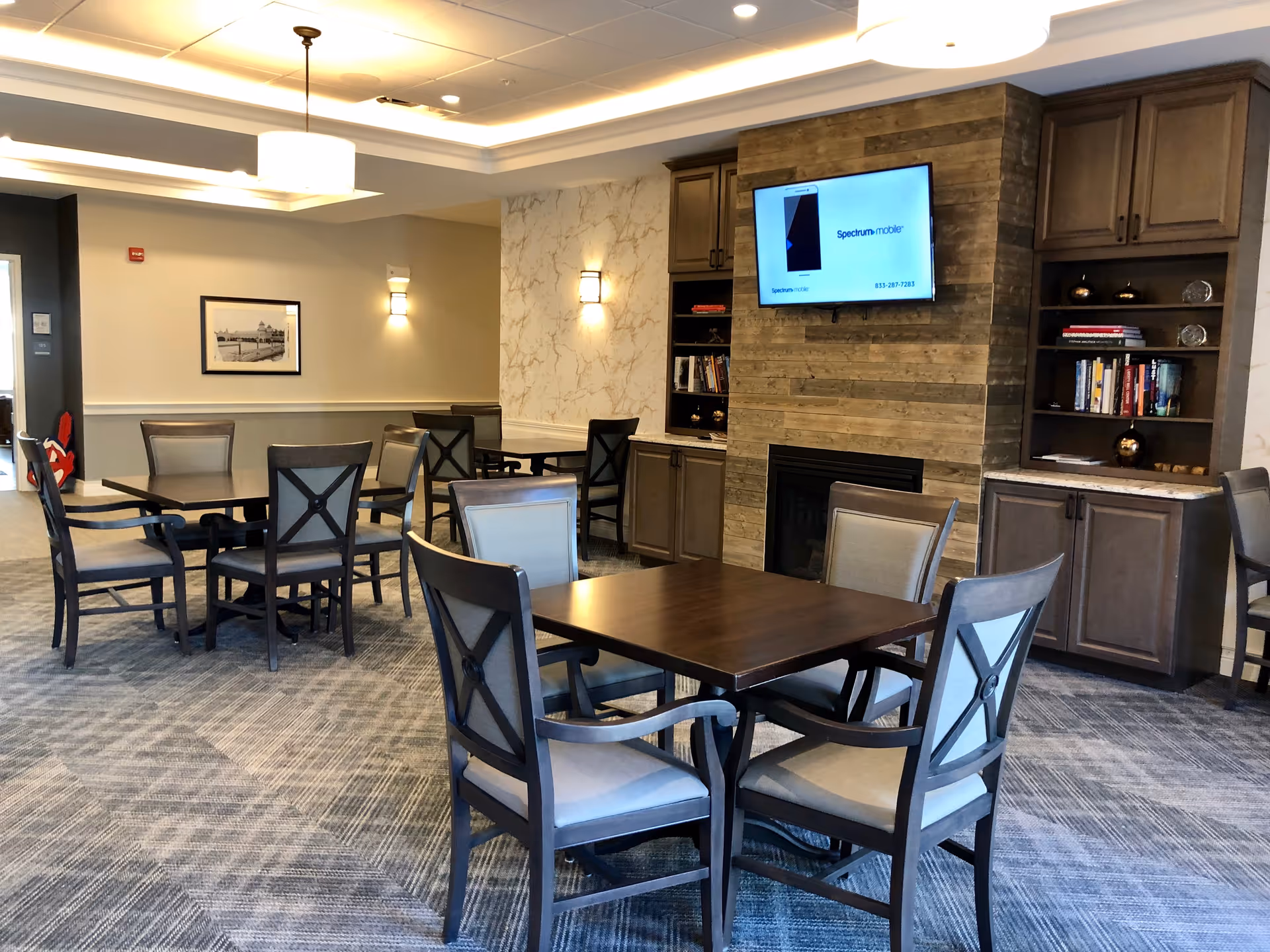 A senior living facility common area with multiple wooden tables and cushioned chairs arranged on a patterned carpet. The room features a fireplace with a wooden panel surround, built-in bookshelves with books and decorative items, and a wall-mounted TV displaying a Spectrum Mobile advertisement. The ceiling has recessed lighting and a hanging light fixture, and the walls are decorated with framed artwork and sconces.