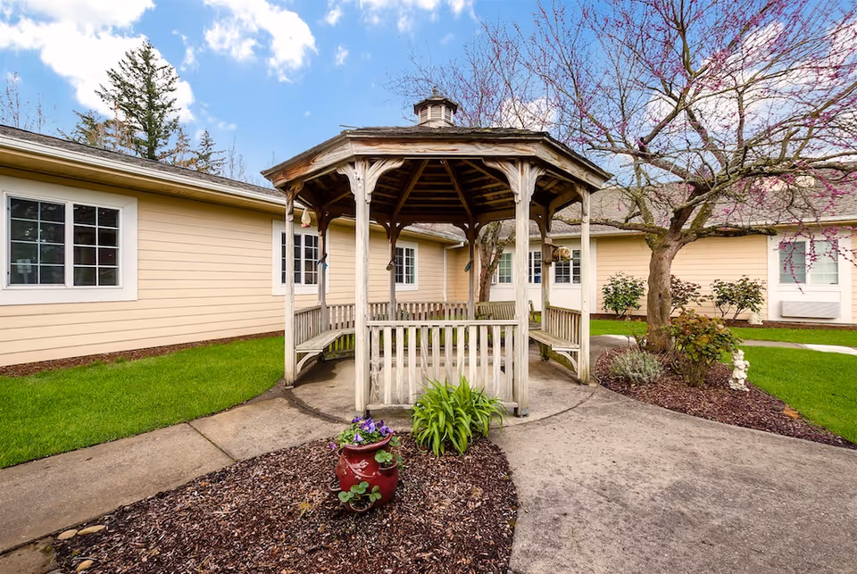 Wooden gazebo in a landscaped courtyard surrounded by single-story beige senior living buildings and flowering trees.