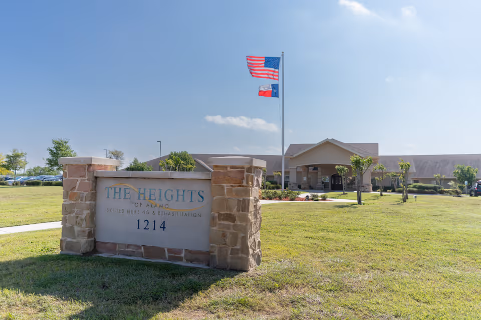Exterior view of The Heights of Alamo skilled nursing and rehabilitation facility with a stone sign in the foreground displaying the facility name and address 1214. The building is visible in the background with a flagpole flying the American and Texas flags. The area is grassy with a few small trees and a clear blue sky.