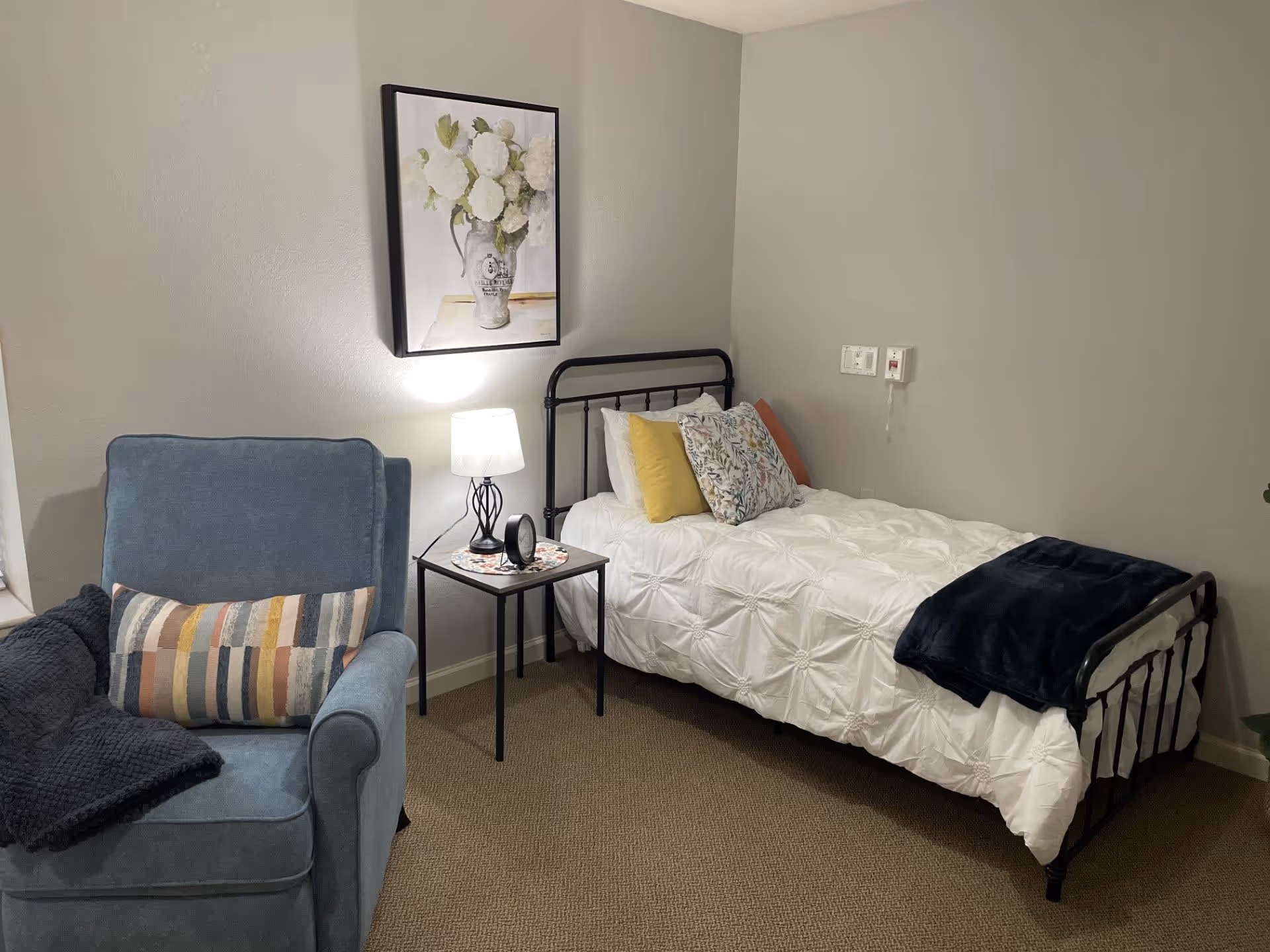 A cozy bedroom with a single black metal bed frame, white comforter, and multiple decorative pillows. Next to the bed is a small black side table with a lamp and a small clock. A blue upholstered armchair with a striped pillow and a dark throw blanket is positioned nearby. A framed picture of white flowers in a vase hangs on the wall above the bed.