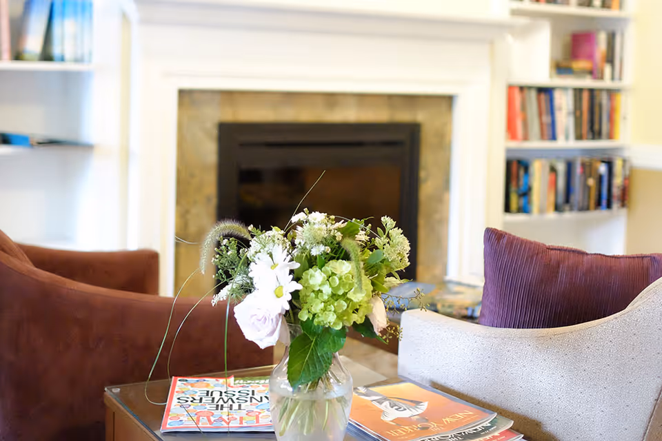 Vase of fresh flowers on a coffee table with magazines in front of a fireplace, surrounded by armchairs and bookshelves.
