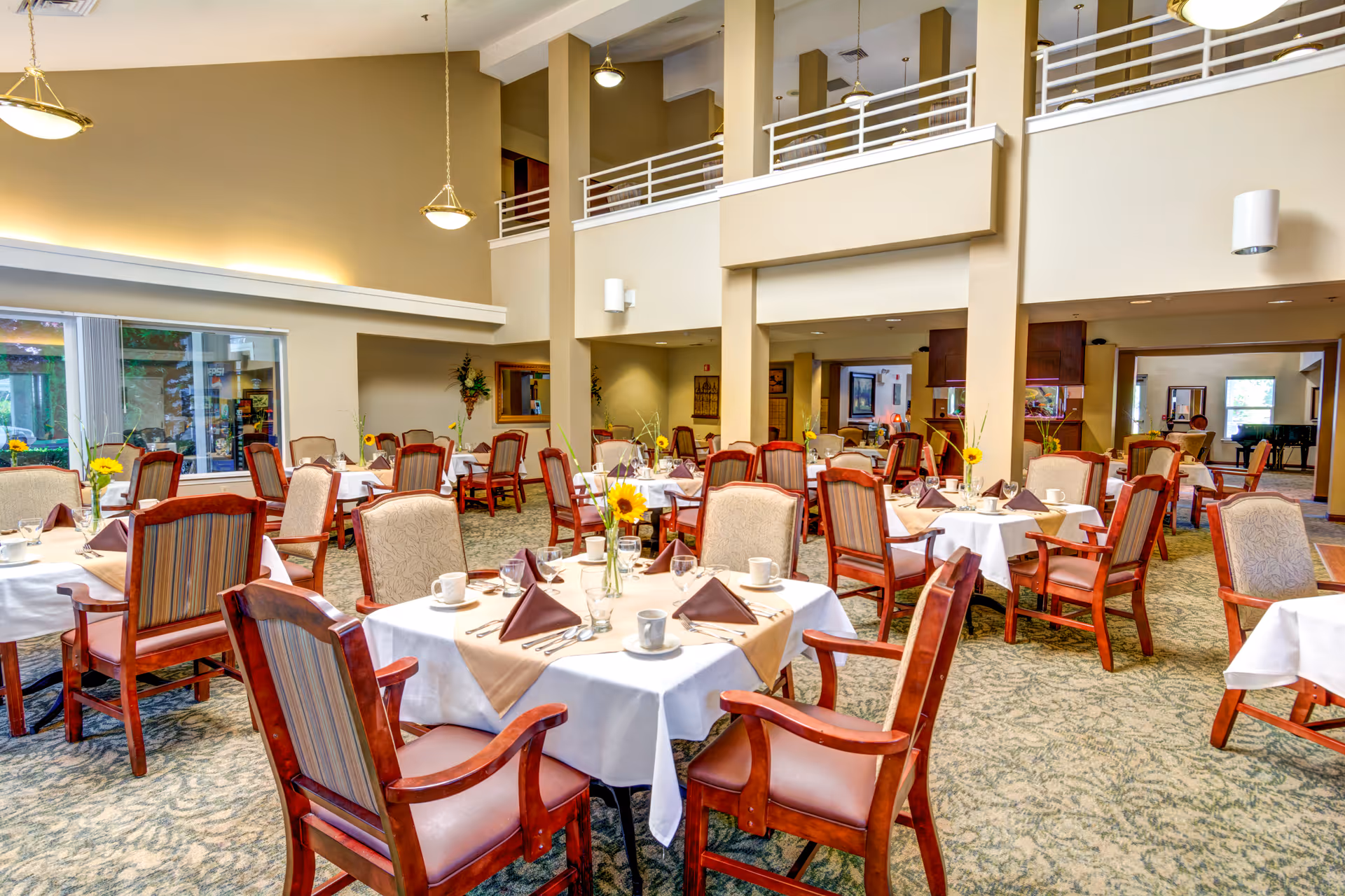 A spacious dining room in an assisted living facility with multiple round tables covered with white tablecloths and beige runners. Each table is set with cups, napkins folded in a triangle, and silverware. The chairs have wooden frames with cushioned seats and backs. The room has high ceilings, large windows, and warm lighting, creating an inviting atmosphere.