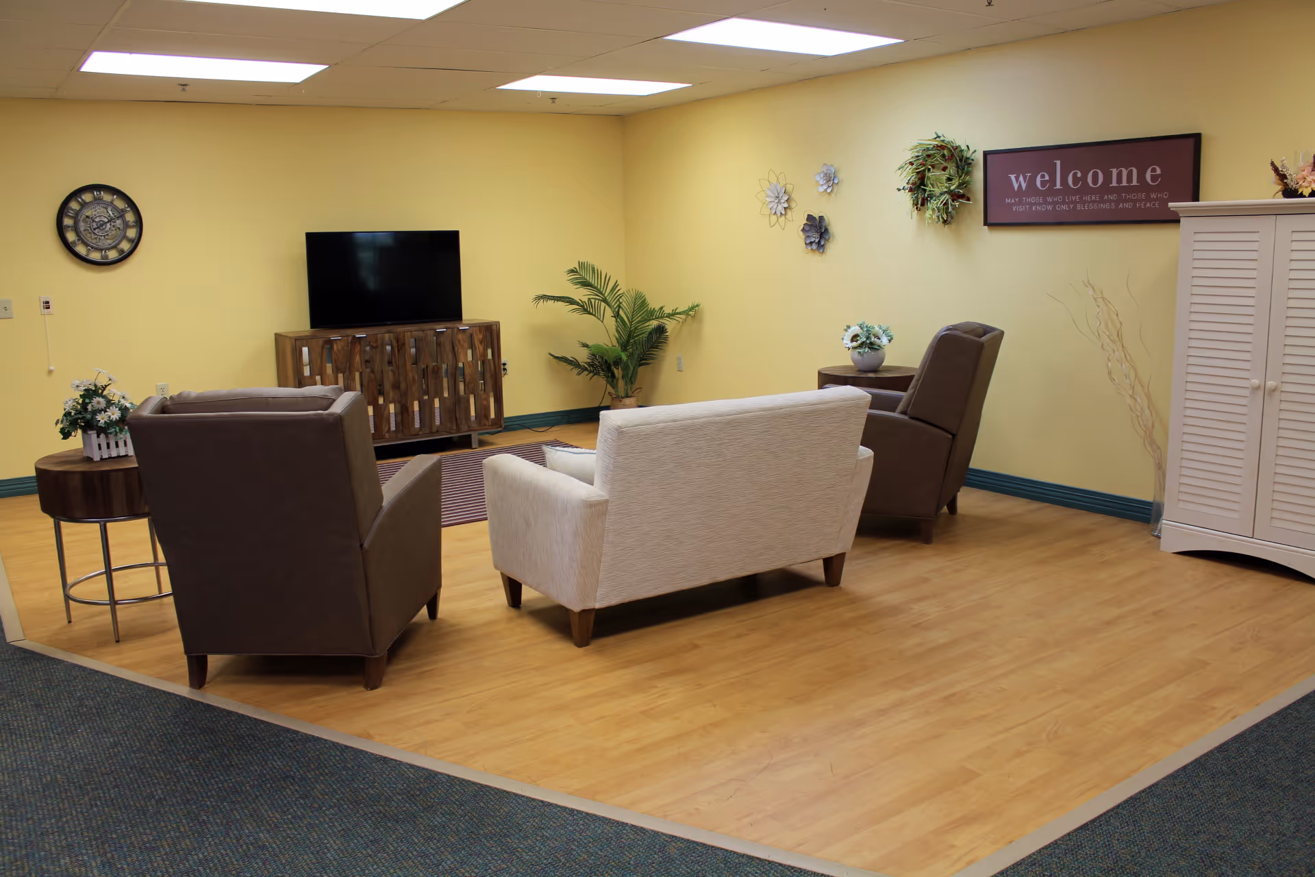 Communal living room with sofas and armchairs arranged around a TV against a yellow wall.