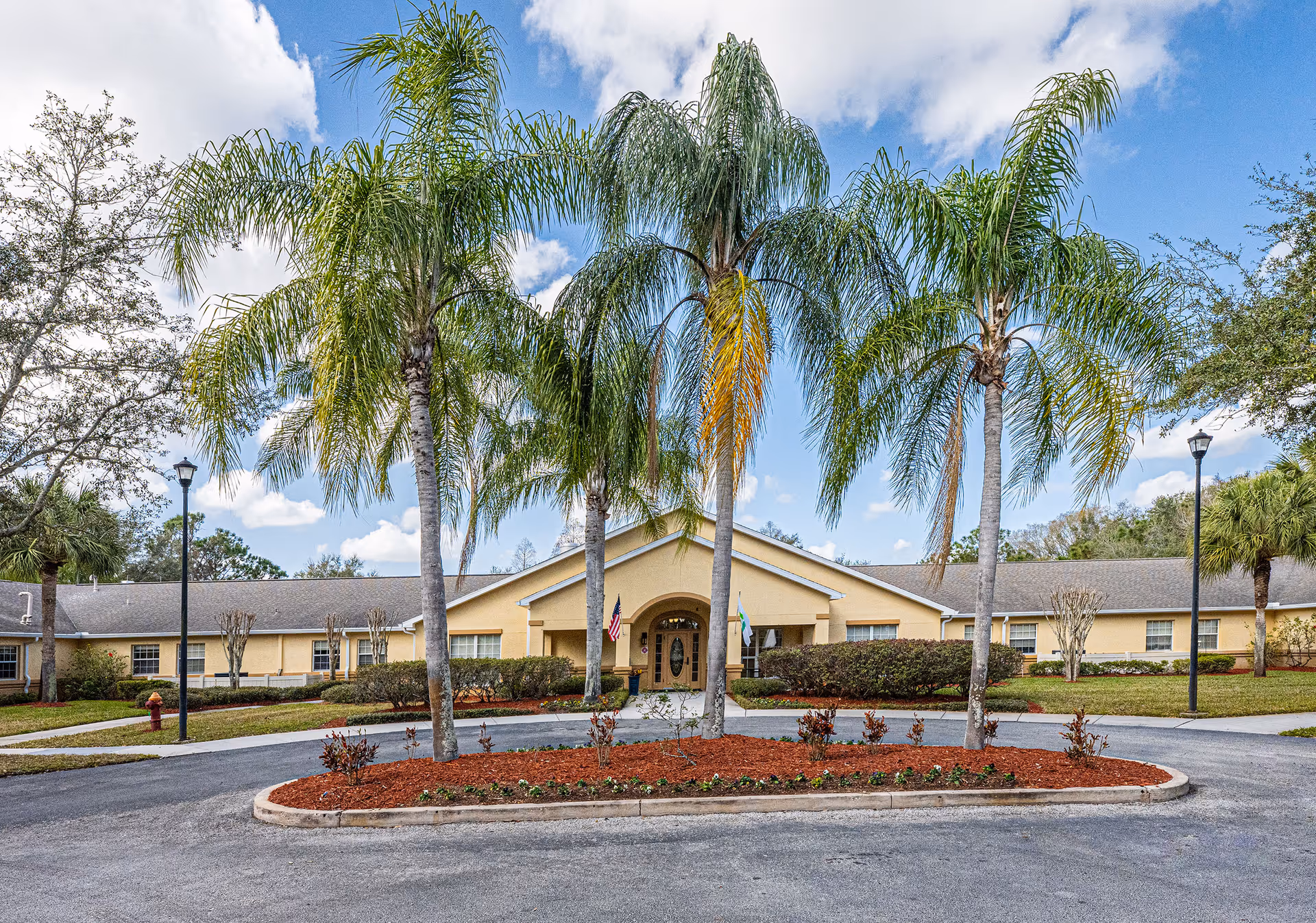 Front entrance of a single-story yellow memory care building framed by palm trees and a circular driveway.